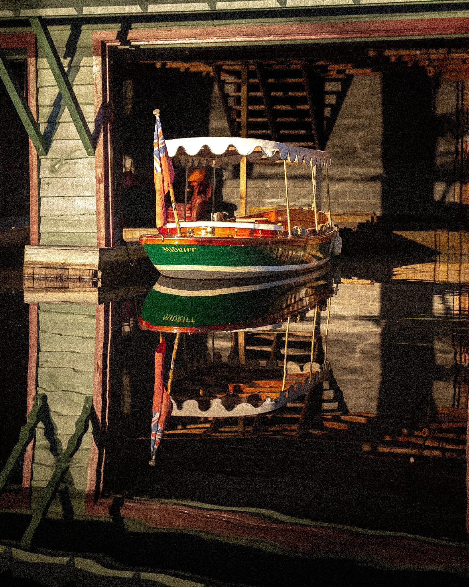 A small wooden boat with a white canopy and a flag on a dock, reflected in the water beneath, with a building and staircase in the background.