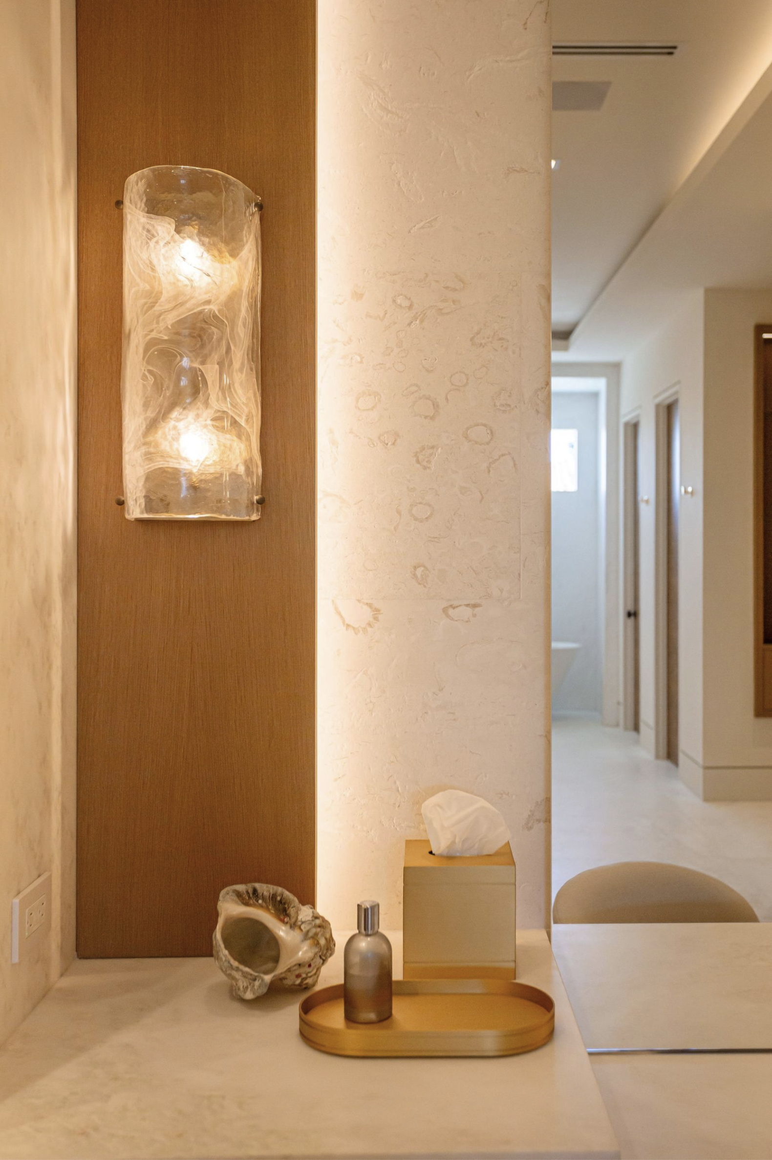 A minimalist bathroom vanity with decor including a large shell, a frosted glass pump dispenser, and a round wooden tray. Behind the vanity, there are textured walls and a hallway with visible doors.