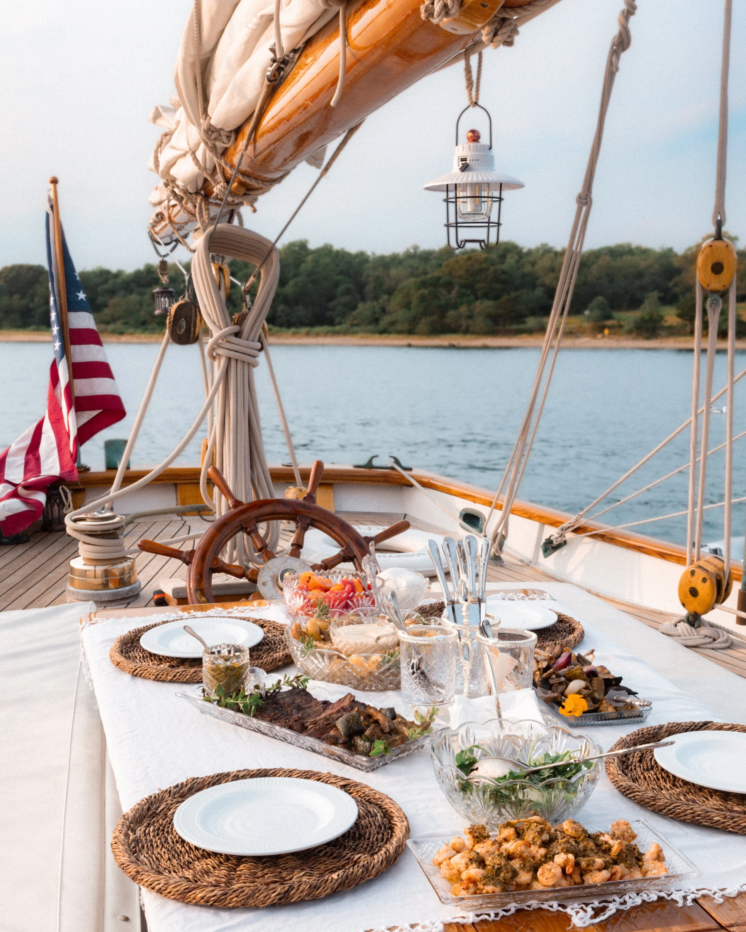 A dining table set up on a boat with various dishes, plates, and glassware, with an outdoor view of water and greenery in the background.