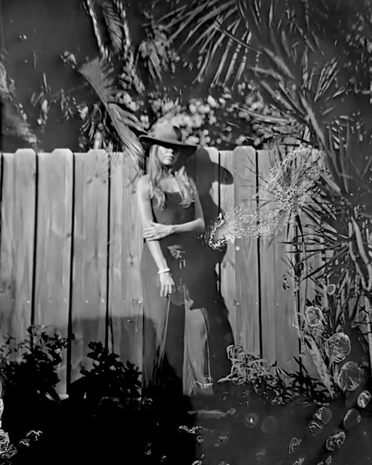 A woman standing outdoors wearing a wide-brimmed hat, black tank top, and black pants, leaning against a wooden fence with lush tropical plants and trees in the background.