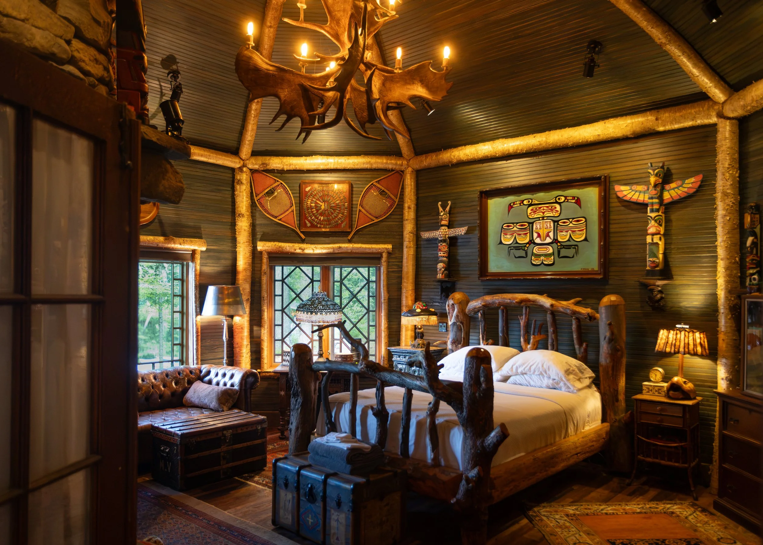 Cozy rustic bedroom with wooden furniture and walls, animal-themed decorations, and warm lighting from a chandelier with animal antler design.