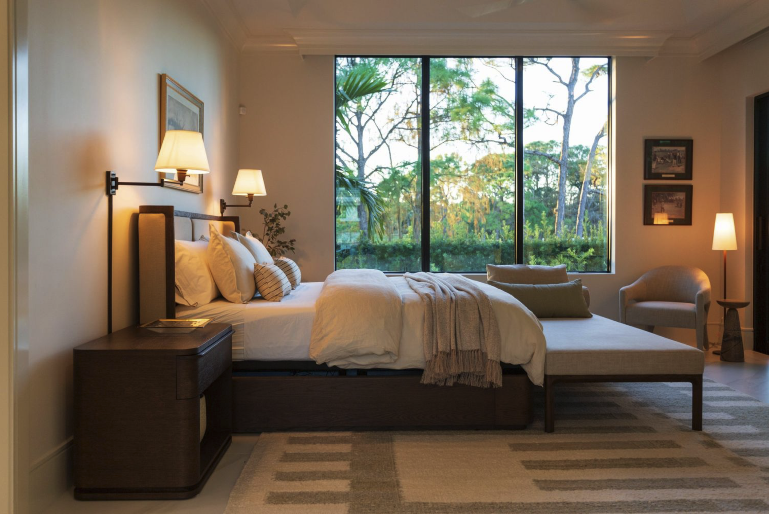 A modern bedroom with a large bed, bedside tables, lamps, and a large window showing trees outside.