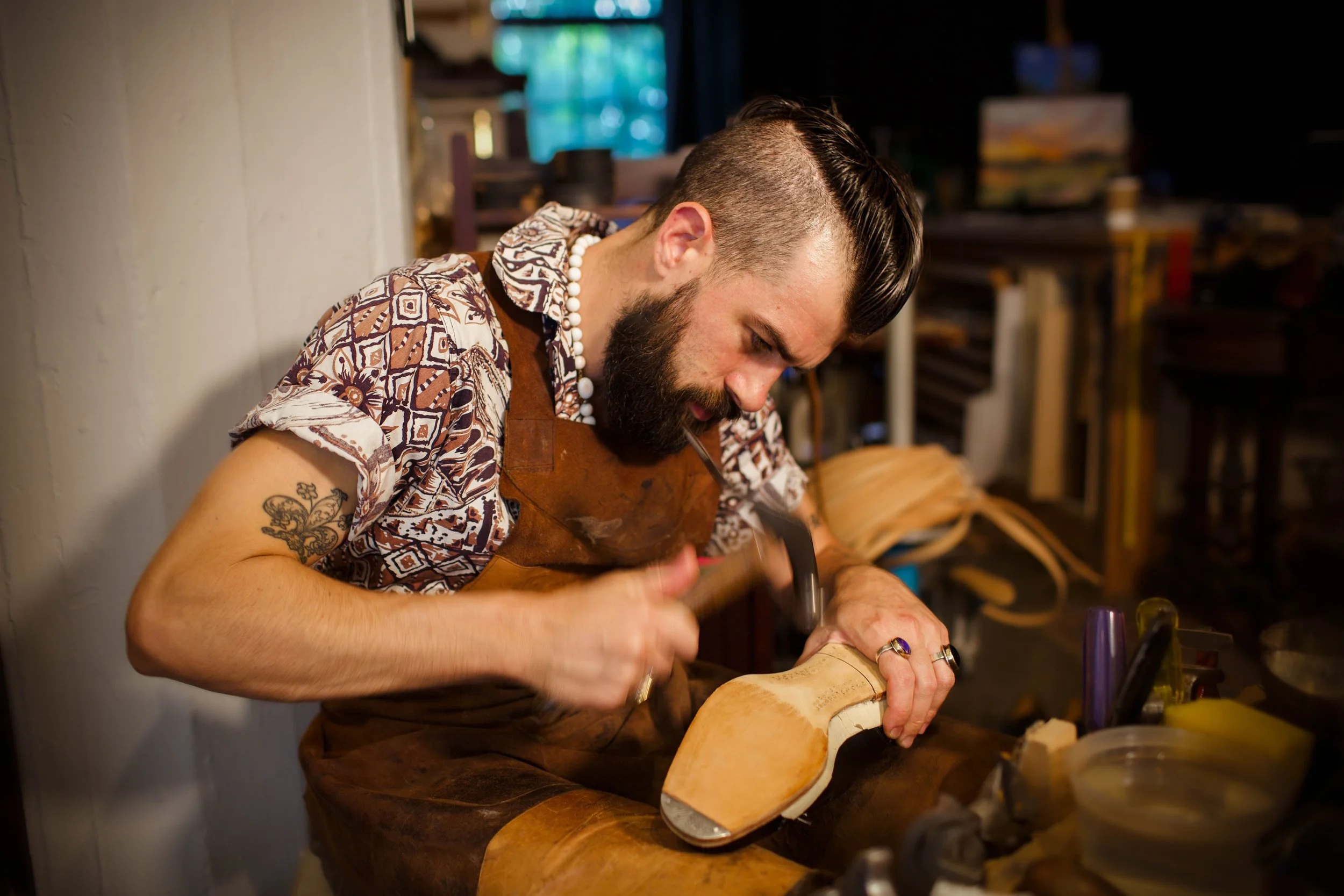 A man with a beard and tattoos working on carving a piece of wood with tools in a workshop.