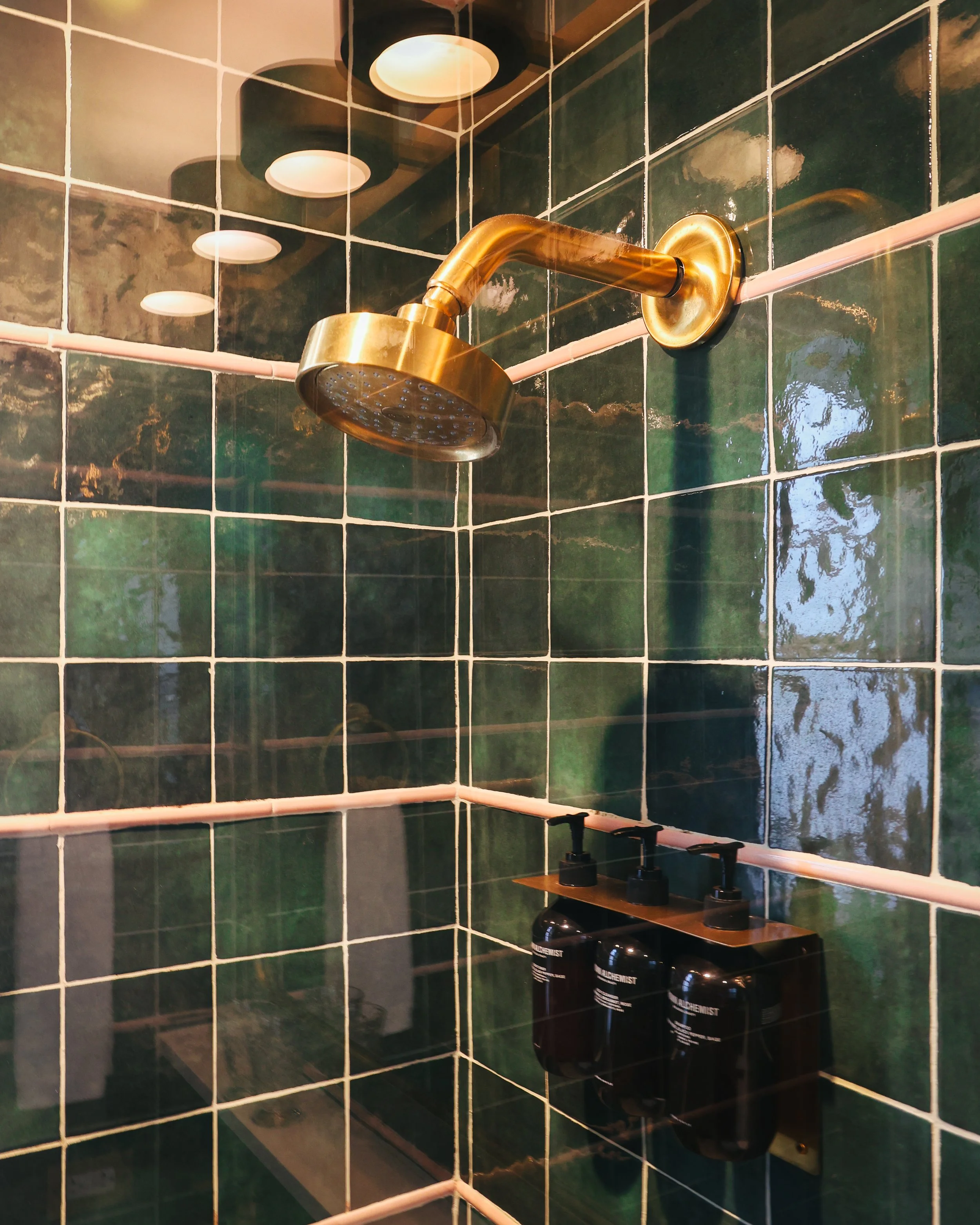 A showerhead mounted on a tiled bathroom wall with green tiles and pink grout, and bottles of hair product below.