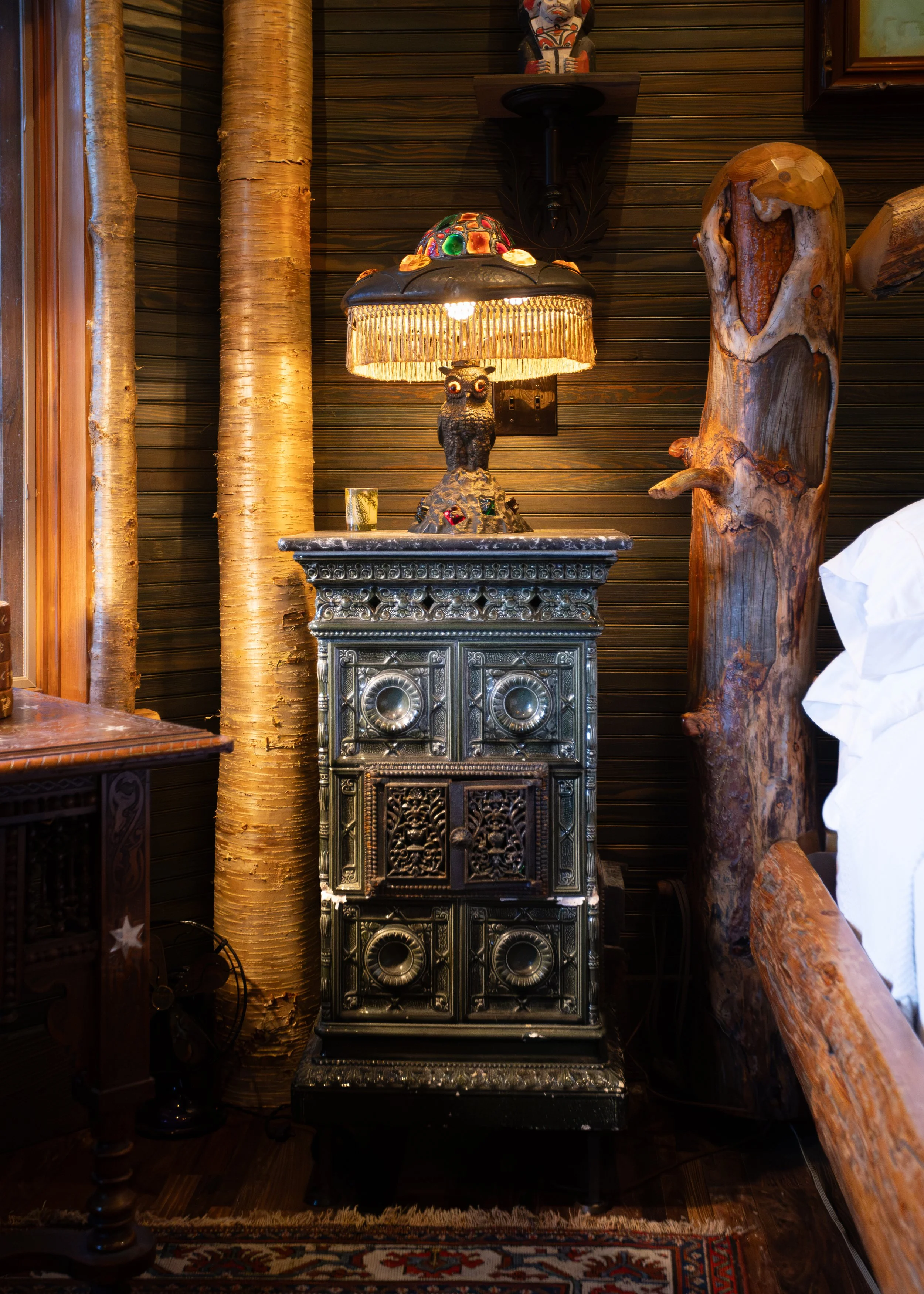 A vintage dark wood side table with ornate metal detailing on the front, topped with a decorative owl-shaped lamp that has a multicolored, mosaic-like shade and fringe, positioned against a dark wood-paneled wall. To the right, there’s a large natural wood sculpture resembling a tree trunk or piece of driftwood. Two tall, light-colored birch logs are standing to the left of the table, and a small portion of a patterned rug with red, white, and blue colors is visible at the bottom.