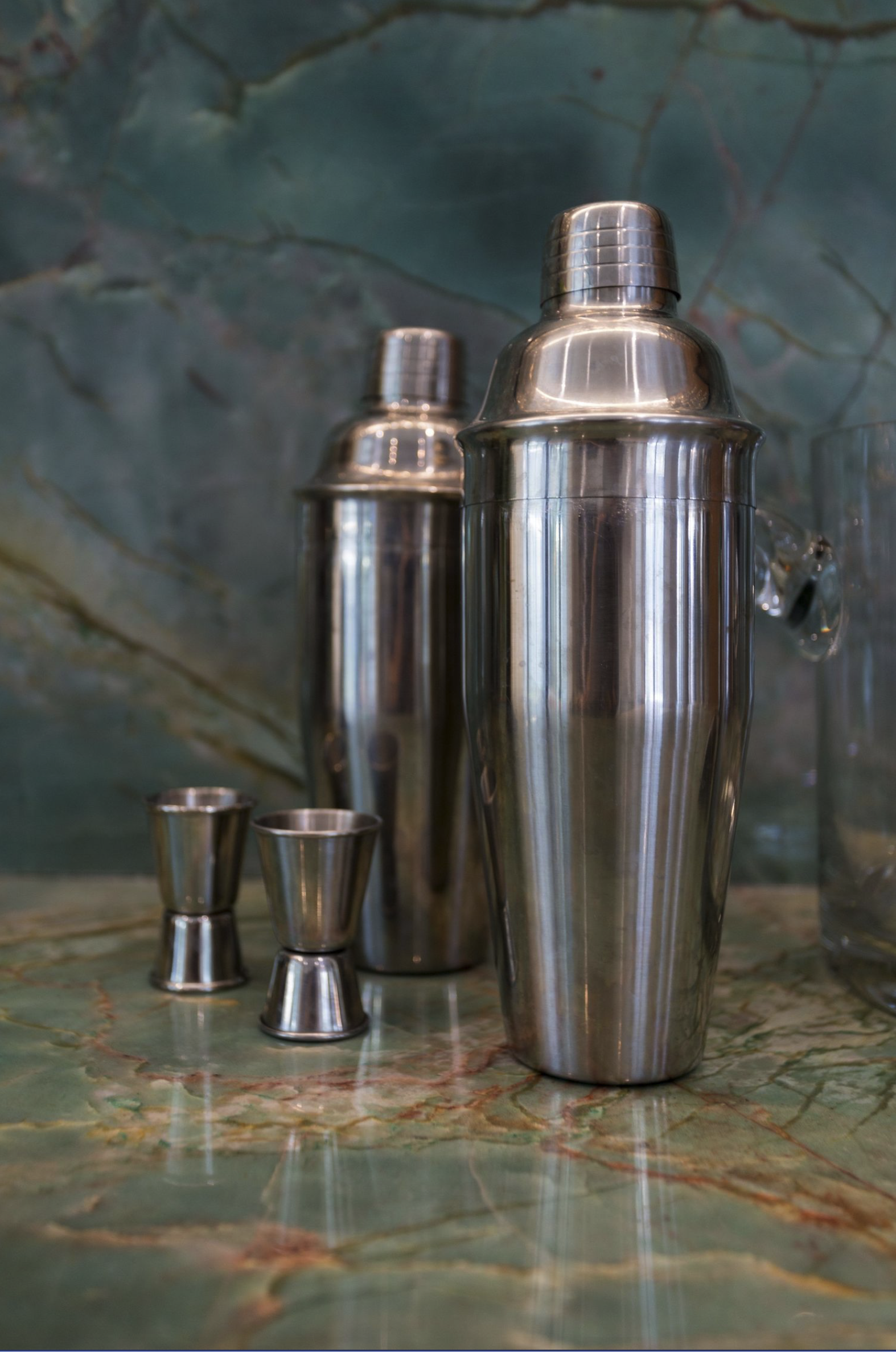 A set of stainless steel cocktail shakers and small cups on a green marble surface with a textured wall in the background.