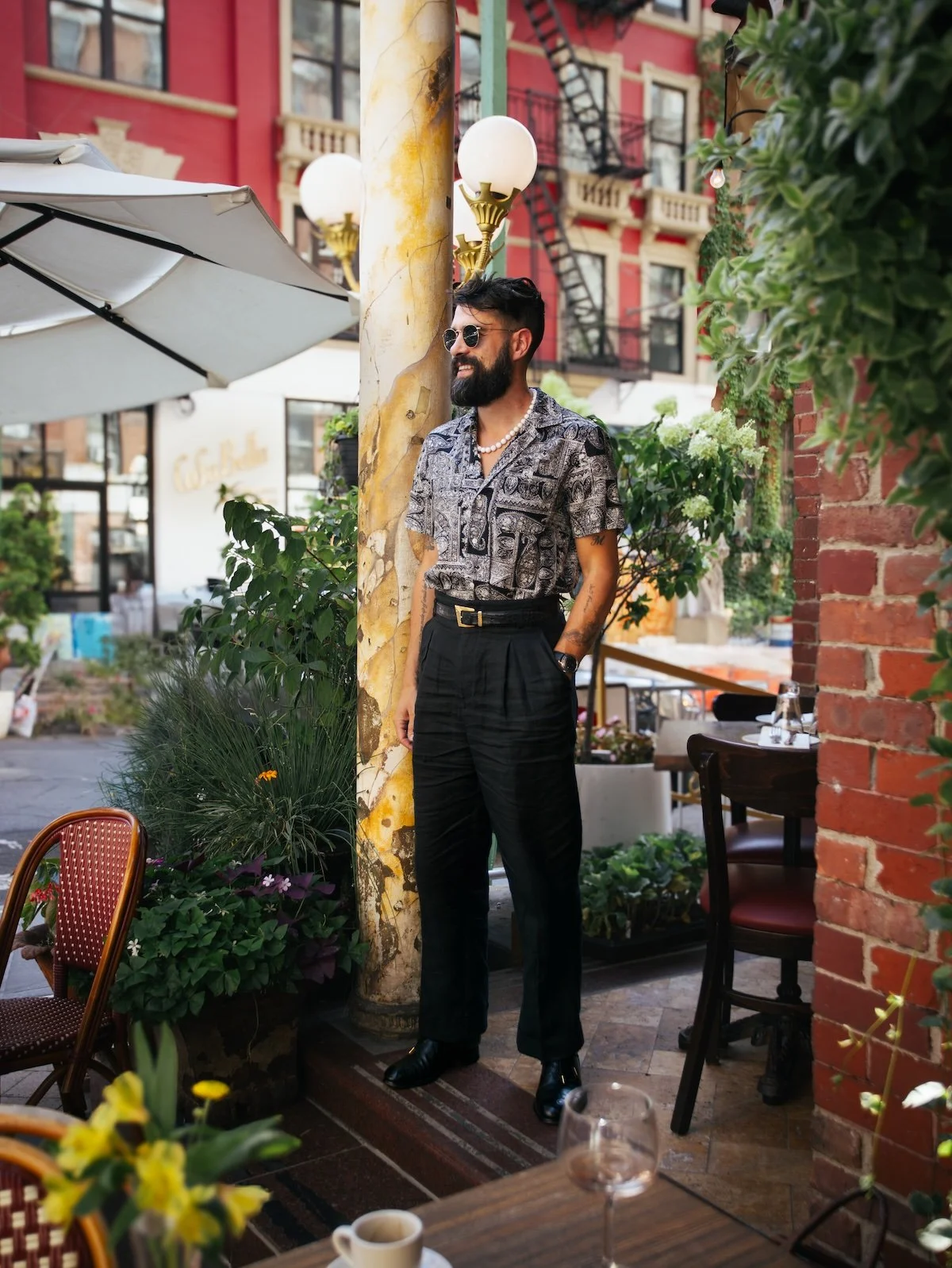 A man with a beard, sunglasses, and a patterned shirt standing outdoors at a cafe or restaurant, leaning against a pole, with greenery and outdoor furniture around him.