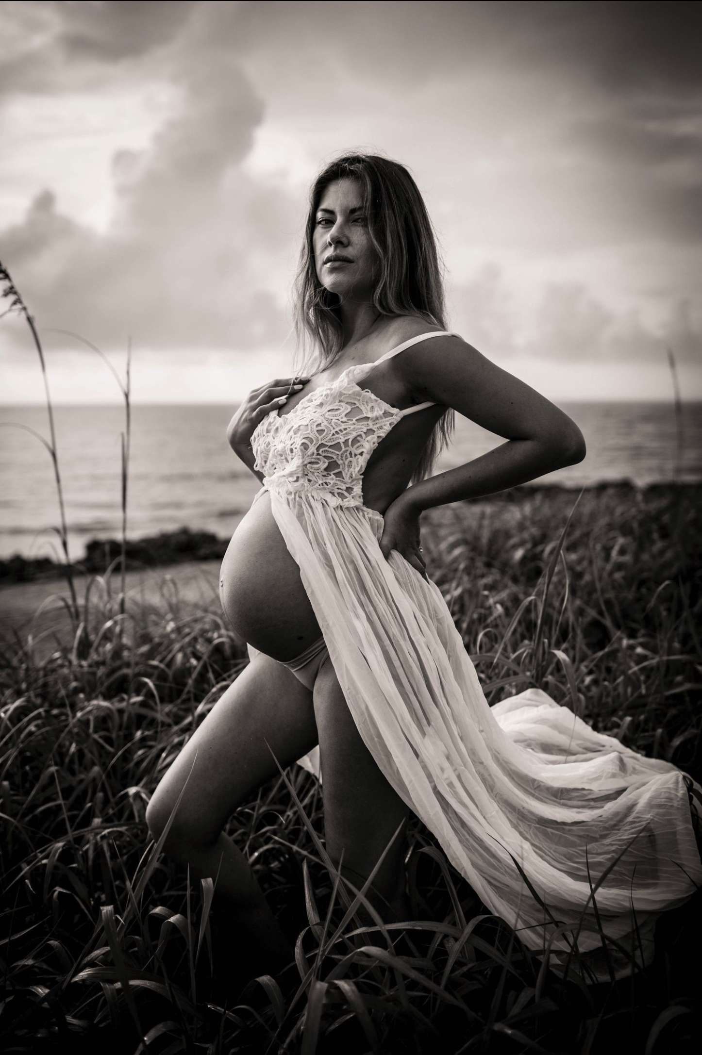 A pregnant woman standing in a grassy field near the ocean, wearing a flowing dress. The photo is in black and white, and she looks at the camera with a serious expression.