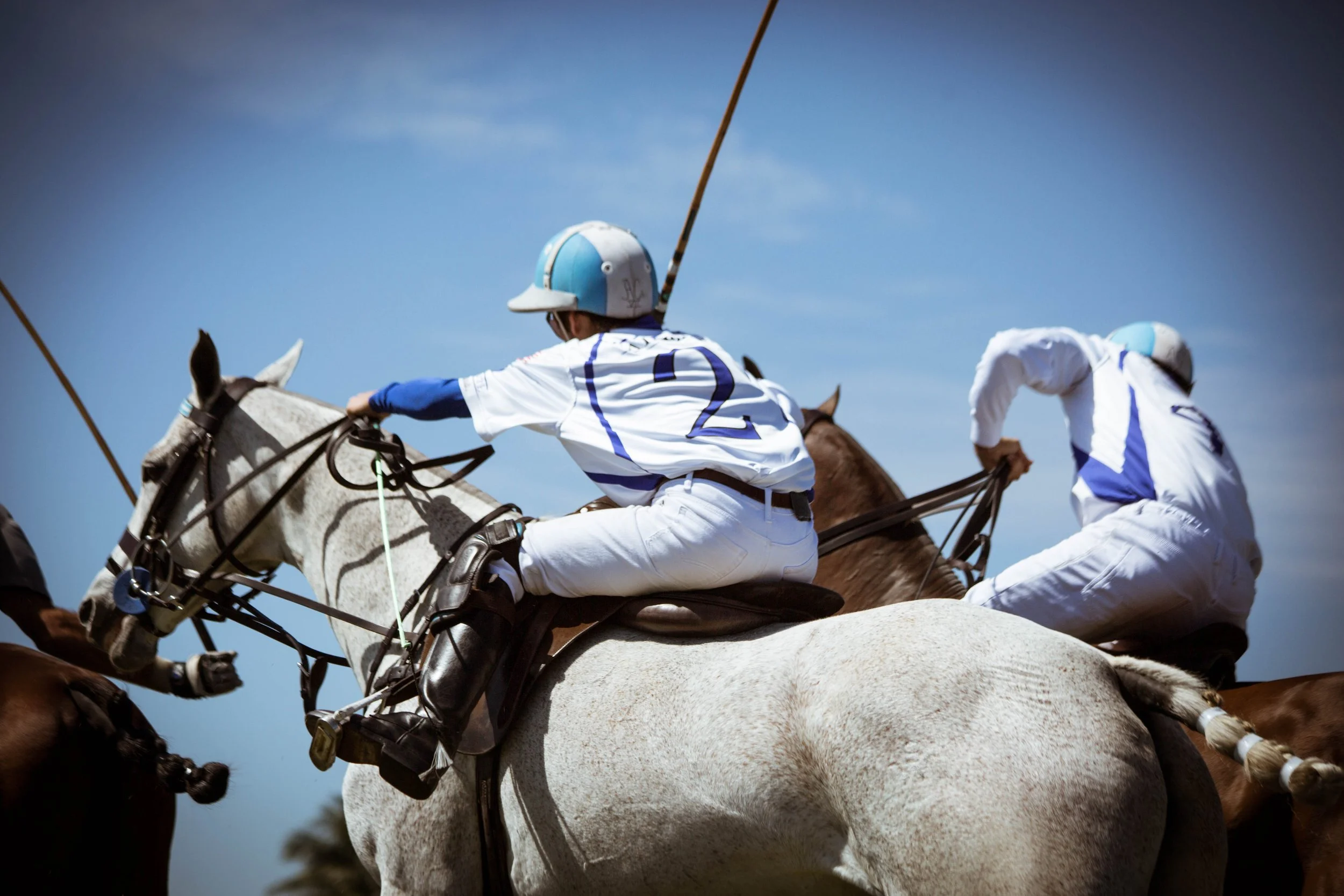 Two polo players in uniform riding horses during a game with a clear blue sky in the background.