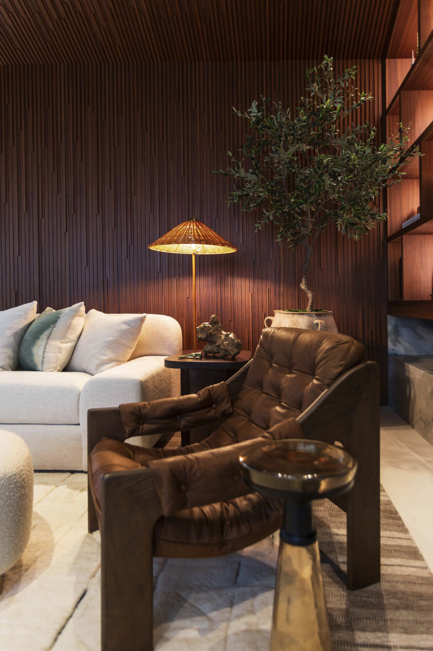 A cozy living room with a beige sofa, brown leather chairs, a wooden side table with decorative items, a wooden wall with a TV shelf, a potted plant, and a warm floor lamp.