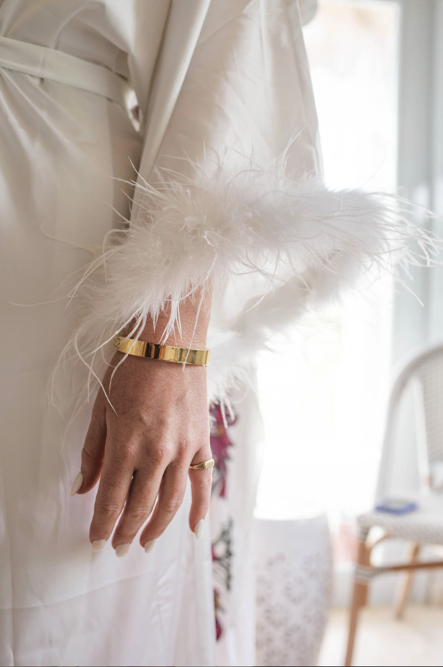 Close-up of a person's hand with a gold ring, wearing a gold bracelet, and white nails, resting on a light-colored fabric with a fluffy white feather accessory attached, near a window with natural light.