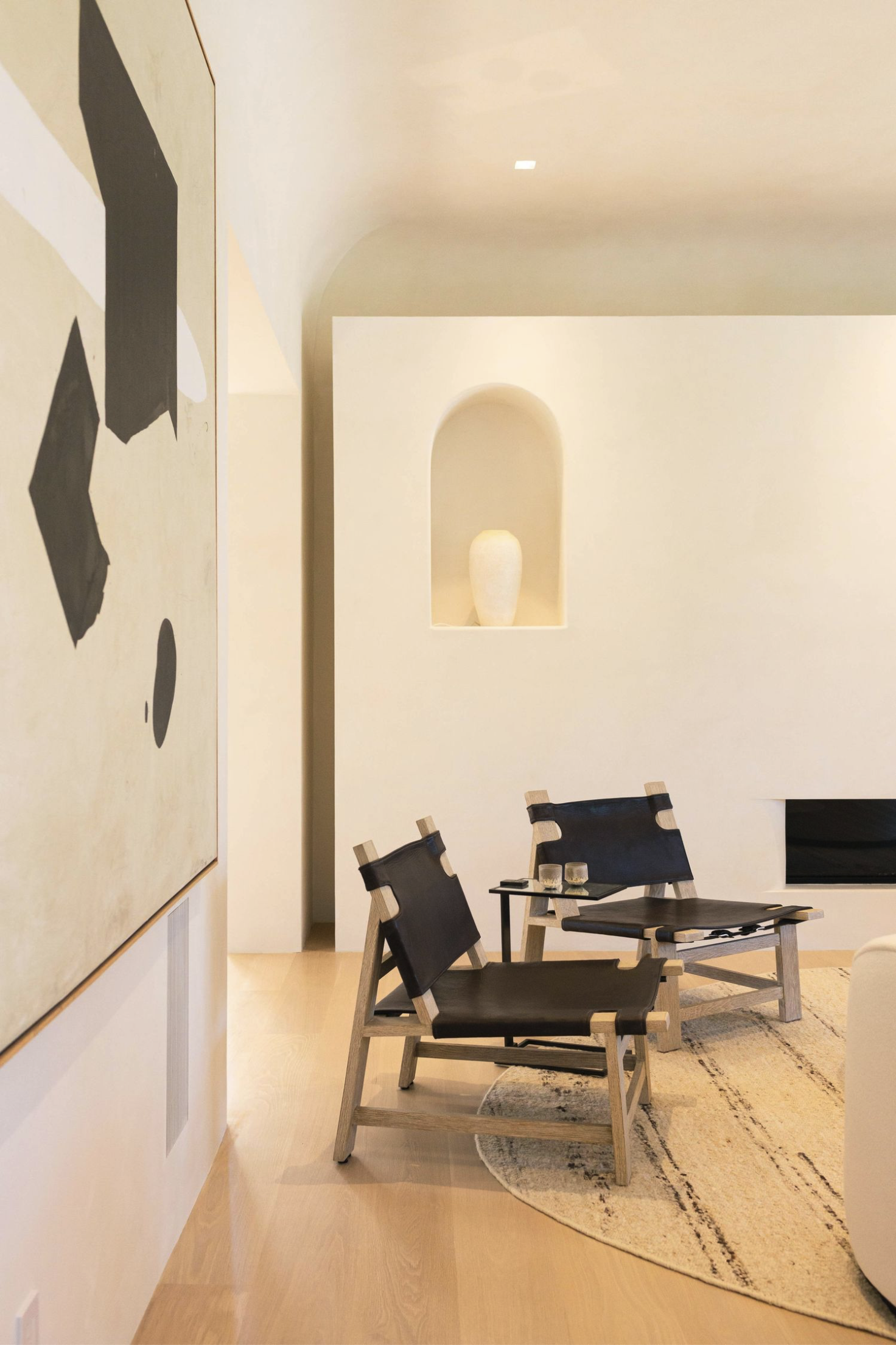 Contemporary living space with four black and natural wood chairs surrounding a small black table on a beige oval rug. A large abstract painting with black, beige, and white shapes hangs on the wall to the left, and a built-in alcove in the wall holds a white decorative vase. Light wood flooring and minimalist design elements create a modern aesthetic.