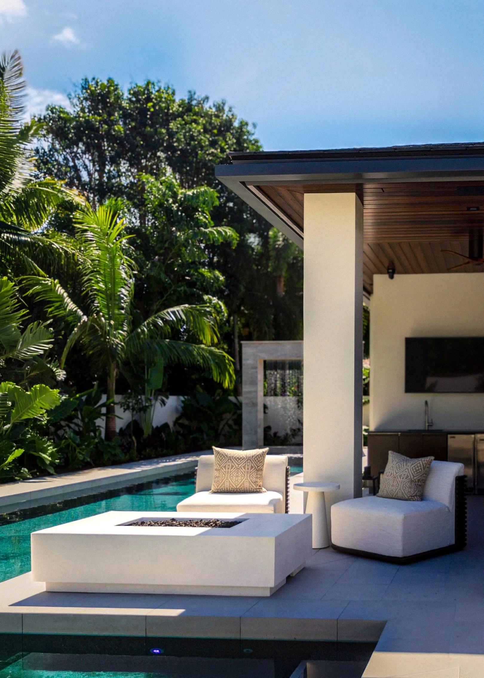 Modern outdoor patio with white lounge chairs, pillows, and a rectangular fire pit near a backyard pool, surrounded by lush green trees and a clear blue sky.