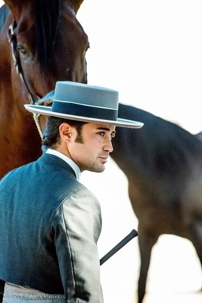 A man in vintage formal attire, including a wide-brimmed hat, stands next to a brown horse.