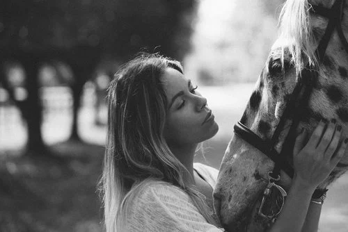 Young woman gently touching the face of a horse in an outdoor setting with trees in the background.