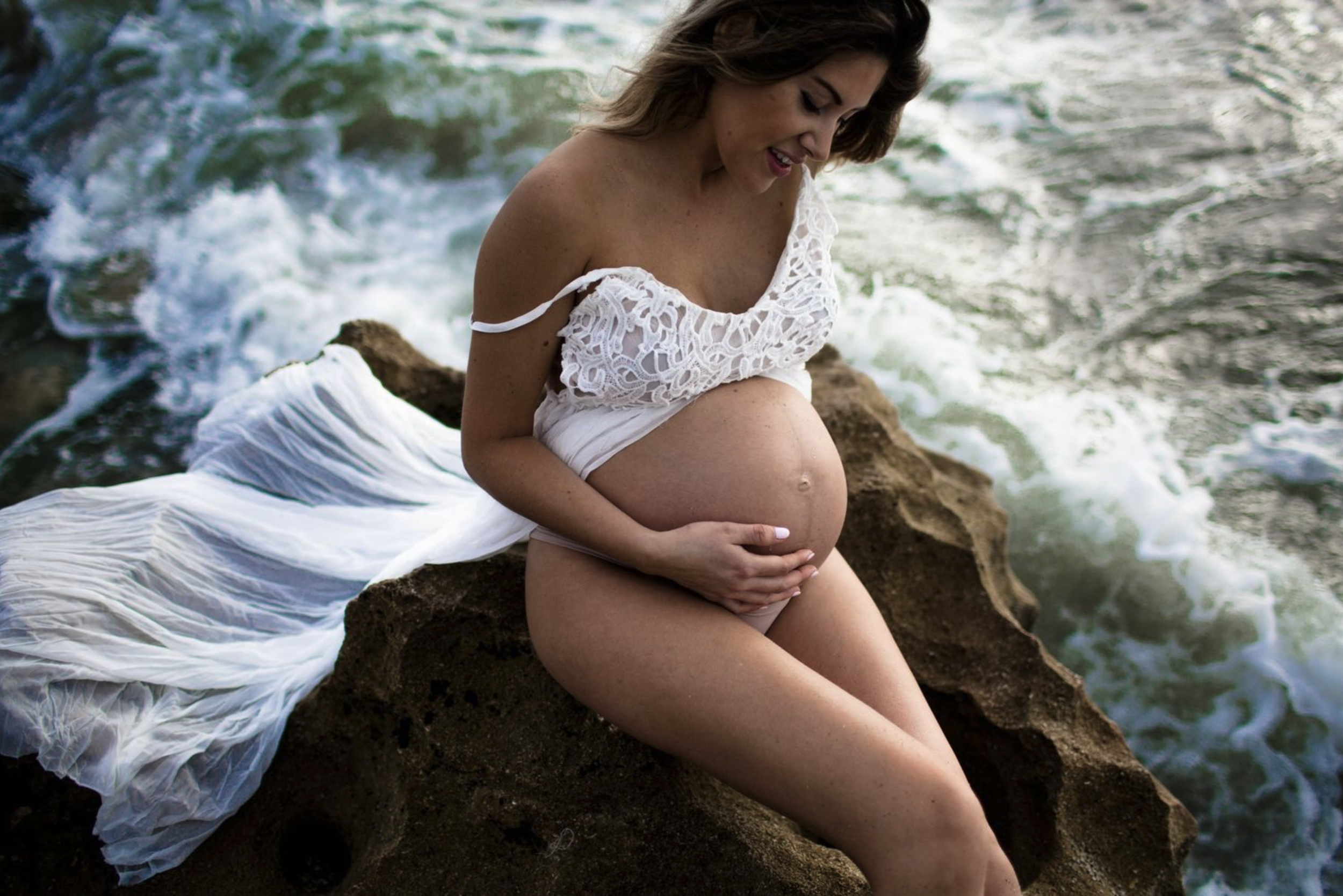 Pregnant woman sitting on rocks by the ocean, smiling, wearing a white lace dress.