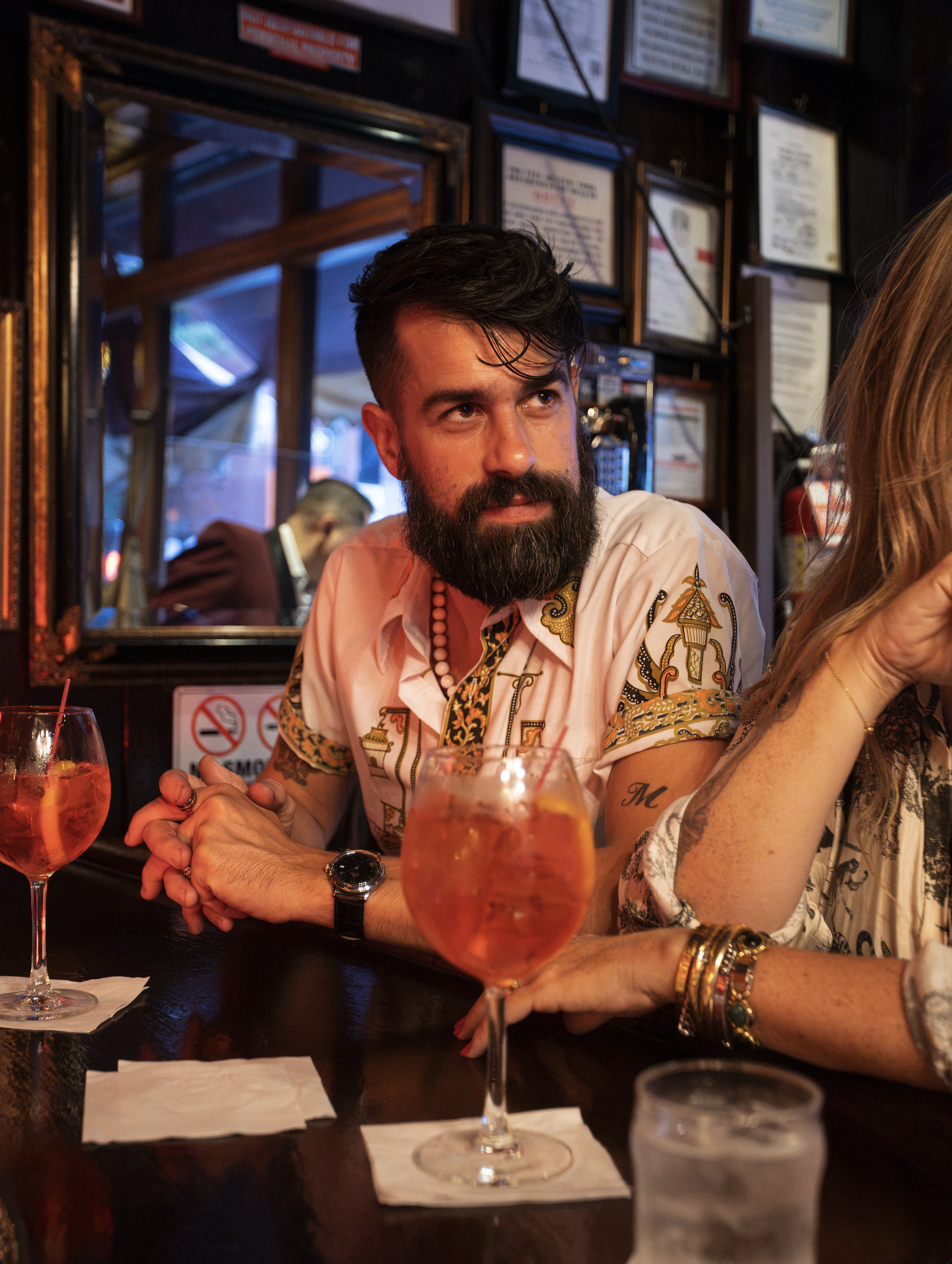 A man with dark hair and a beard sitting at a bar table in a restaurant, wearing a light-colored shirt with colorful embroidery. He has tattoos on his arms, a watch, and several bracelets on his wrists. Two glasses with pink cocktails are in front of him. Part of a woman is visible to his right. The background shows framed certificates or awards on a dark wall and a bartender working behind the bar.