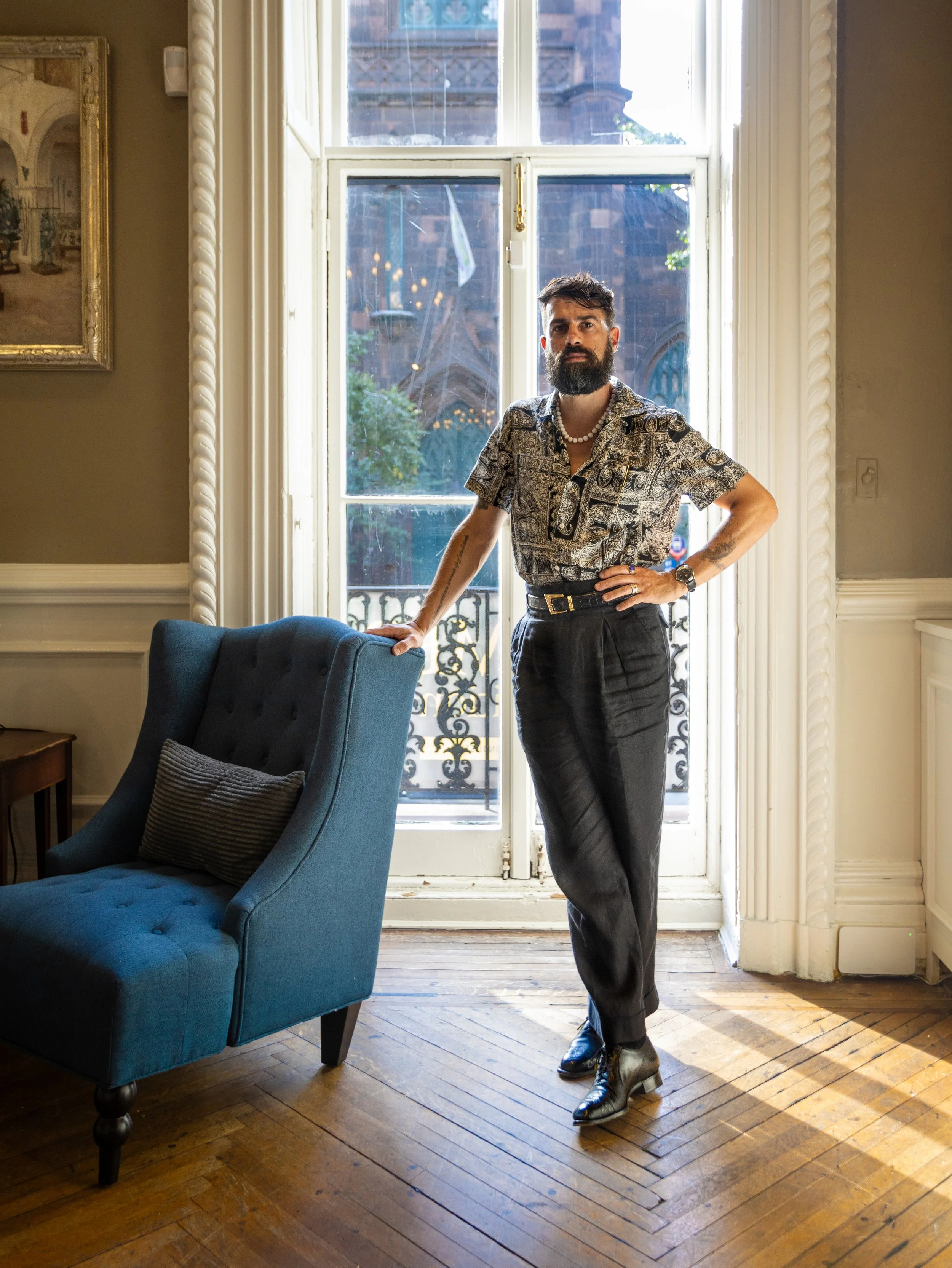 A man with a beard and styled hair, wearing a patterned shirt, black trousers, and shiny shoes, stands inside a room with wood flooring near a large window. He has his left hand on his hip and his right hand on a blue upholstered chair.