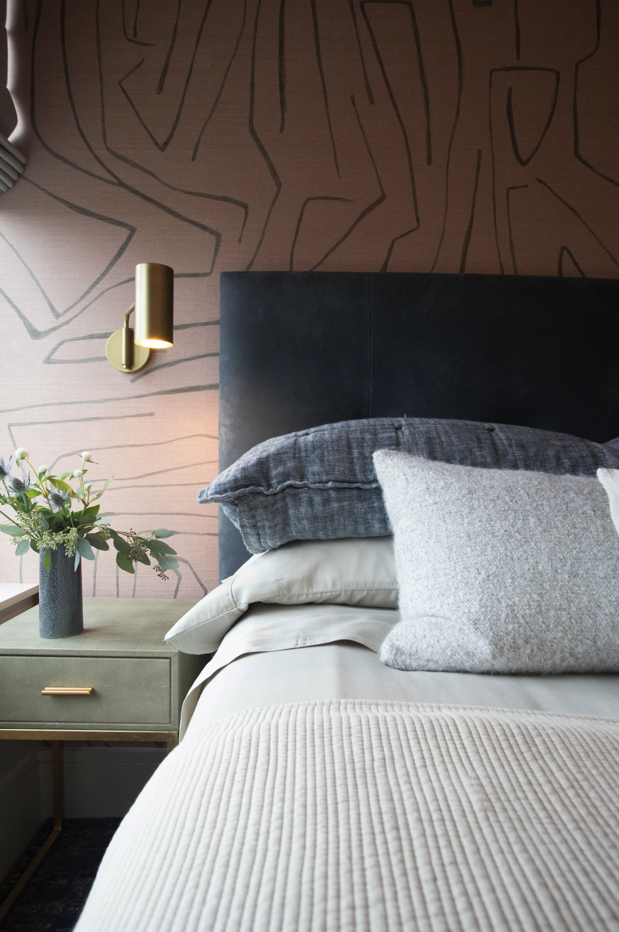 Close-up of a neatly made bed with a gray headboard, layered pillows including a dark gray one and a textured light gray one, beige sheets, and a beige quilted bedspread. Beside the bed, there is a green nightstand with a modern gray vase holding a bouquet of flowers, and a gold wall-mounted reading light on a pink wall with an abstract black line pattern.