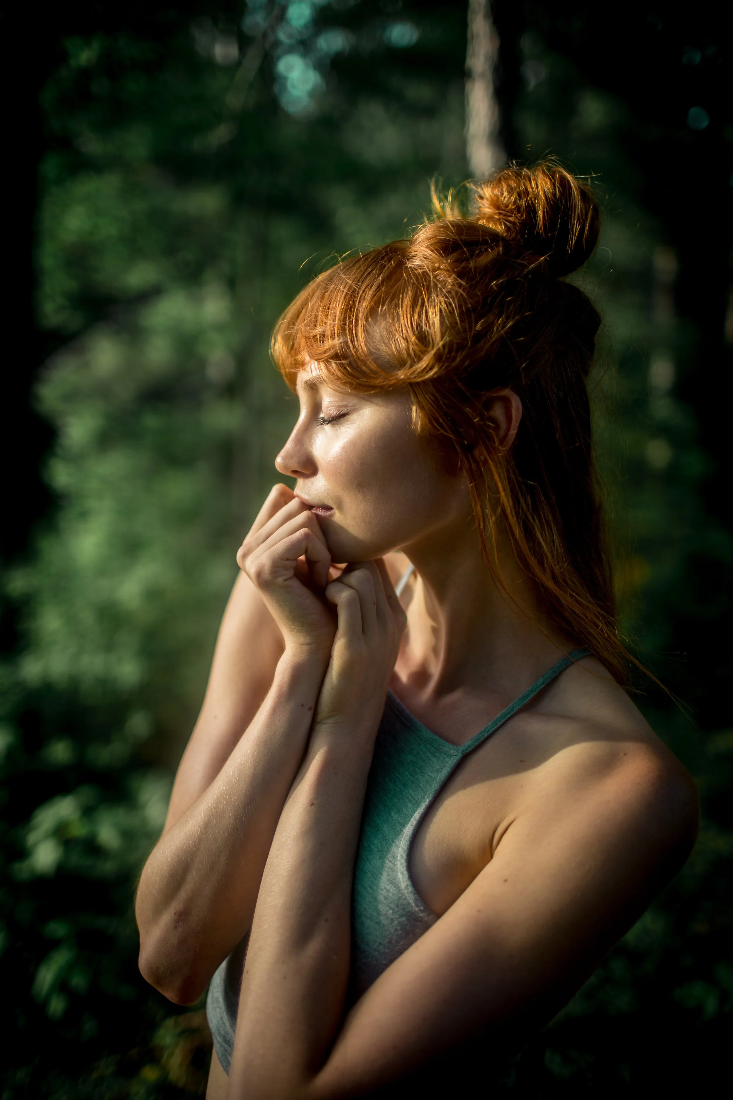 A young woman with red hair styled in a bun, with her eyes closed and her hand near her chin, standing outdoors in a green, sunlit forest.