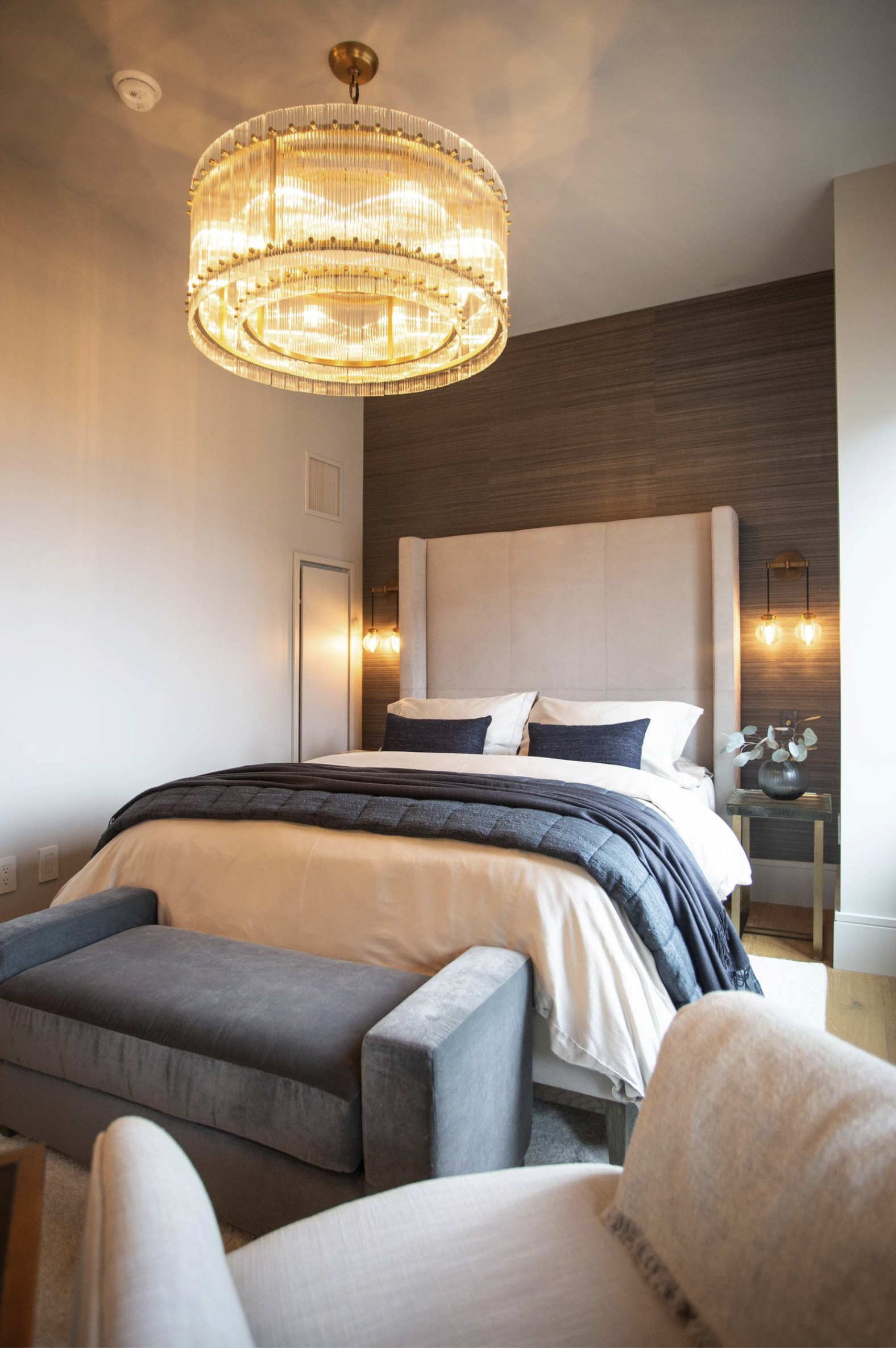 A cozy bedroom with a large bed that has beige and dark gray bedding, with a denim bench at its foot, a beige upholstered headboard, modern wall lamps, and a chandelier hanging from the ceiling, with cream-colored walls and a wood-paneled accent wall.