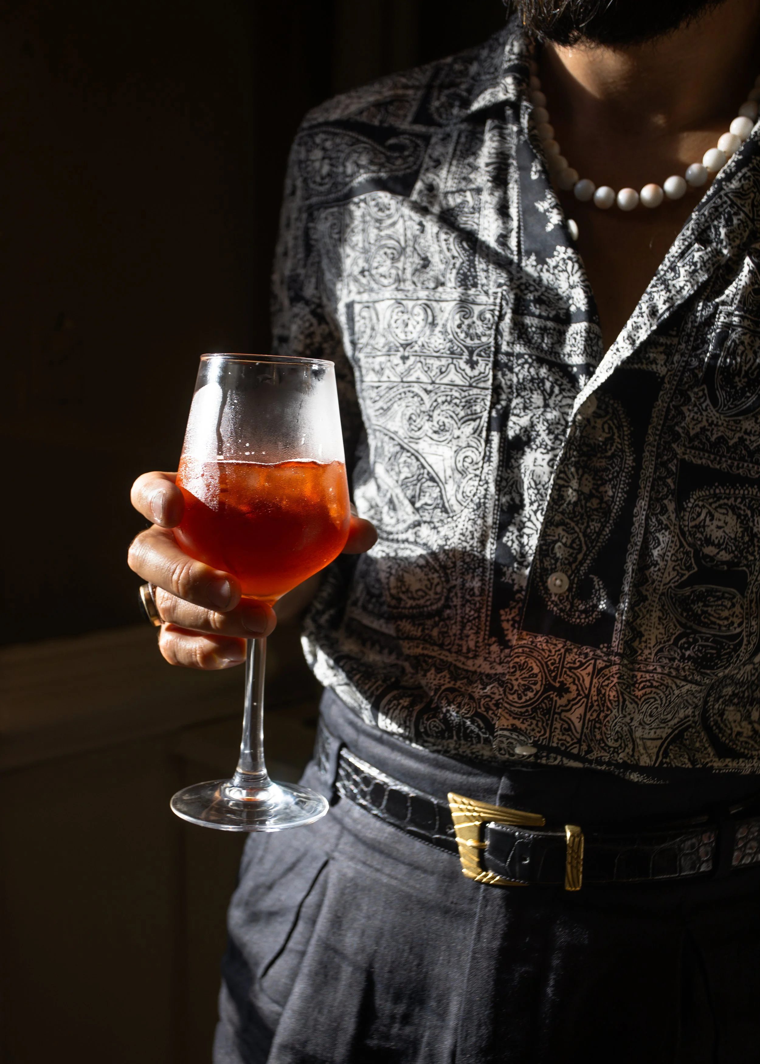Person holding a glass of red wine, wearing a patterned shirt, with a pearl necklace and a gold belt buckle visible.