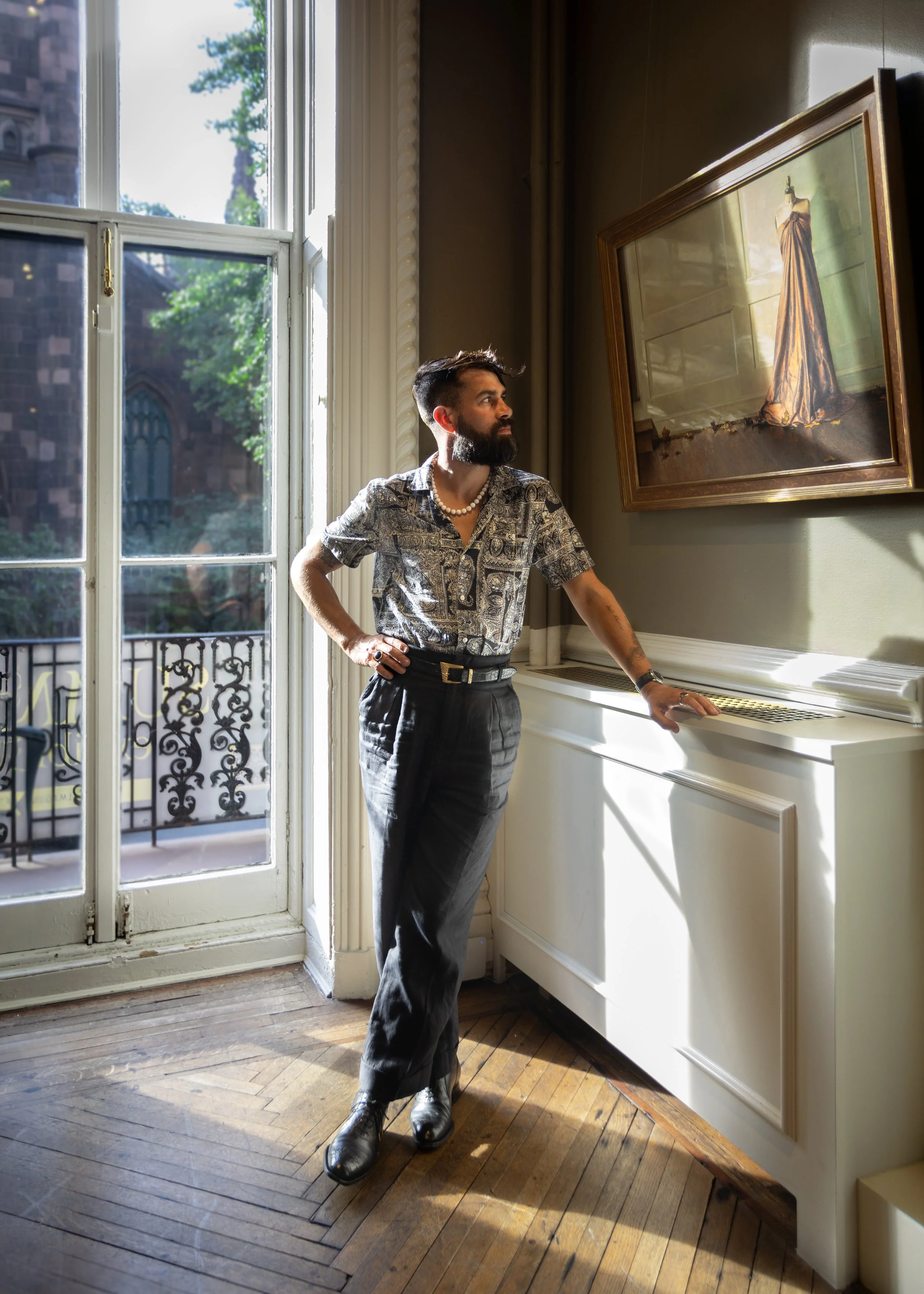 A man with a beard, dressed in vintage-style clothing, stands in an elegant room with a large window, wooden floor, and framed artwork. He wears a patterned shirt, pearl necklace, and black pants, with one hand on his hip and the other resting on a white piano.