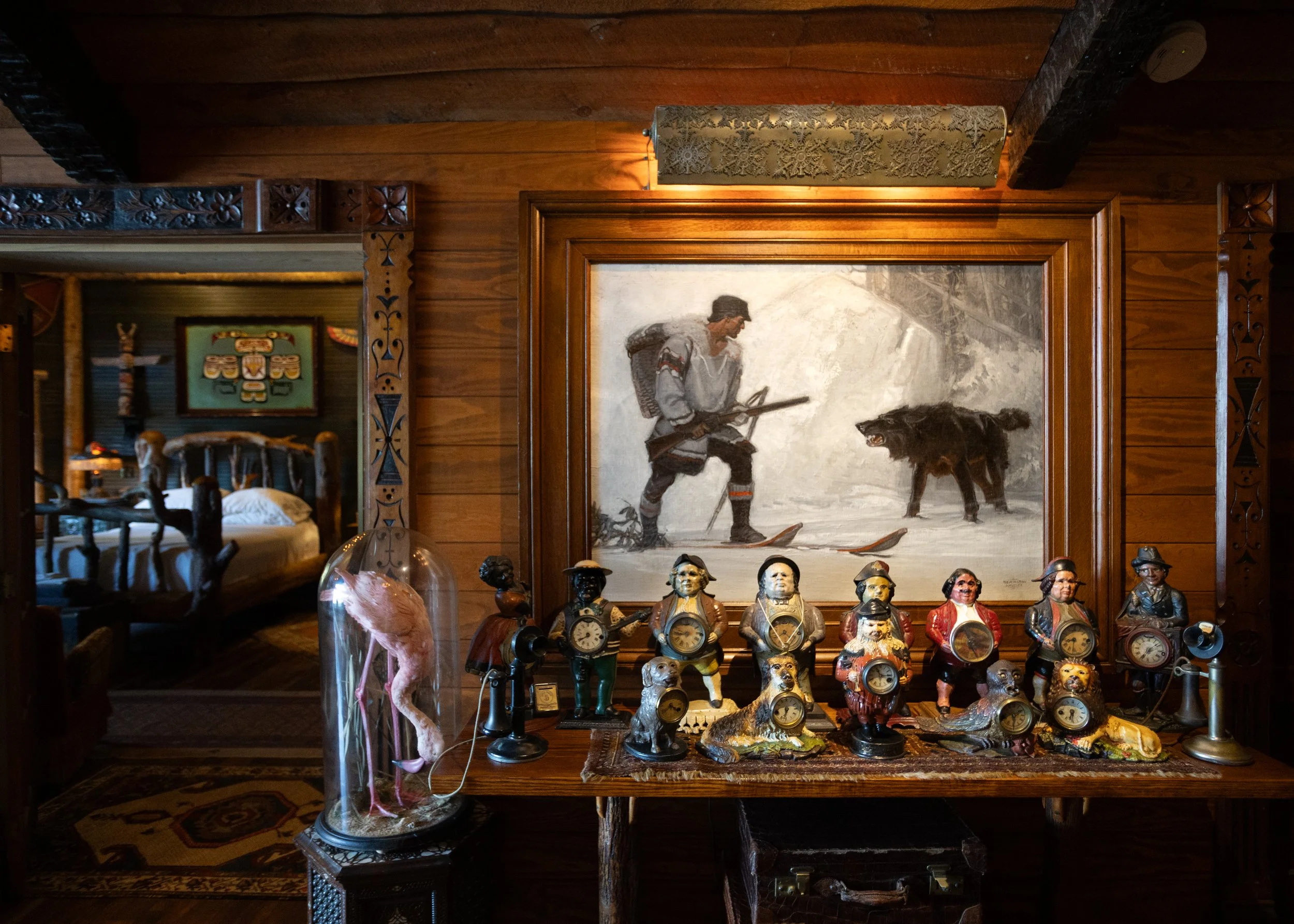 A wooden room decorated with Native American art and artifacts. The front features small figurines with clocks, and in the background, there's a large painting of a man with a dog. The room has warm lighting and intricate carved wood details.