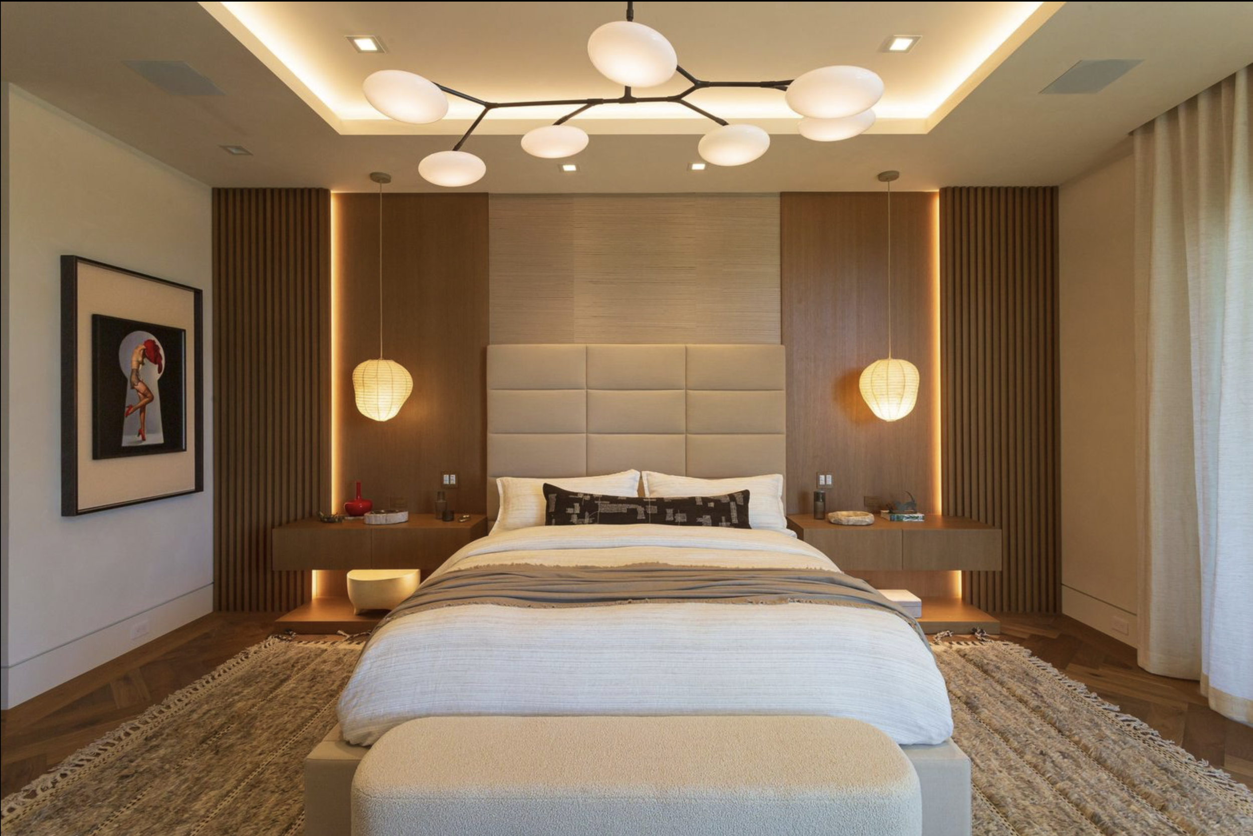 A modern bedroom with a centered bed, wooden side tables, hanging lamps, a framed art piece, and a ceiling light fixture.