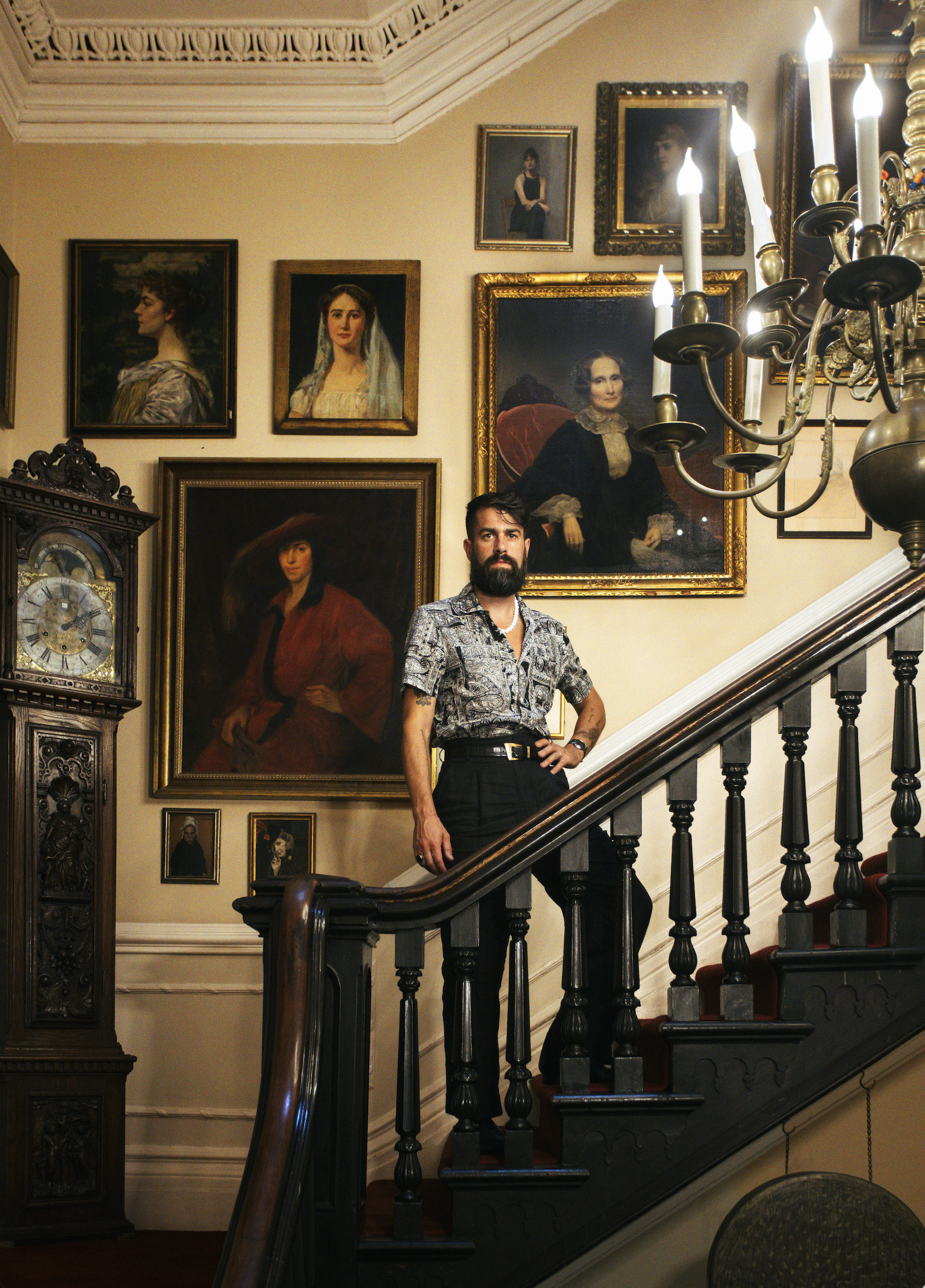 A man with a beard and dark hair, wearing a patterned shirt and black pants, stands on a staircase in a room decorated with framed classic and modern portrait paintings. The staircase has a dark wooden handrail with black balusters. There is an ornate grandfather clock to the left and a chandelier with lit candles hanging above. The room has beige walls, crown molding, and various framed artworks.