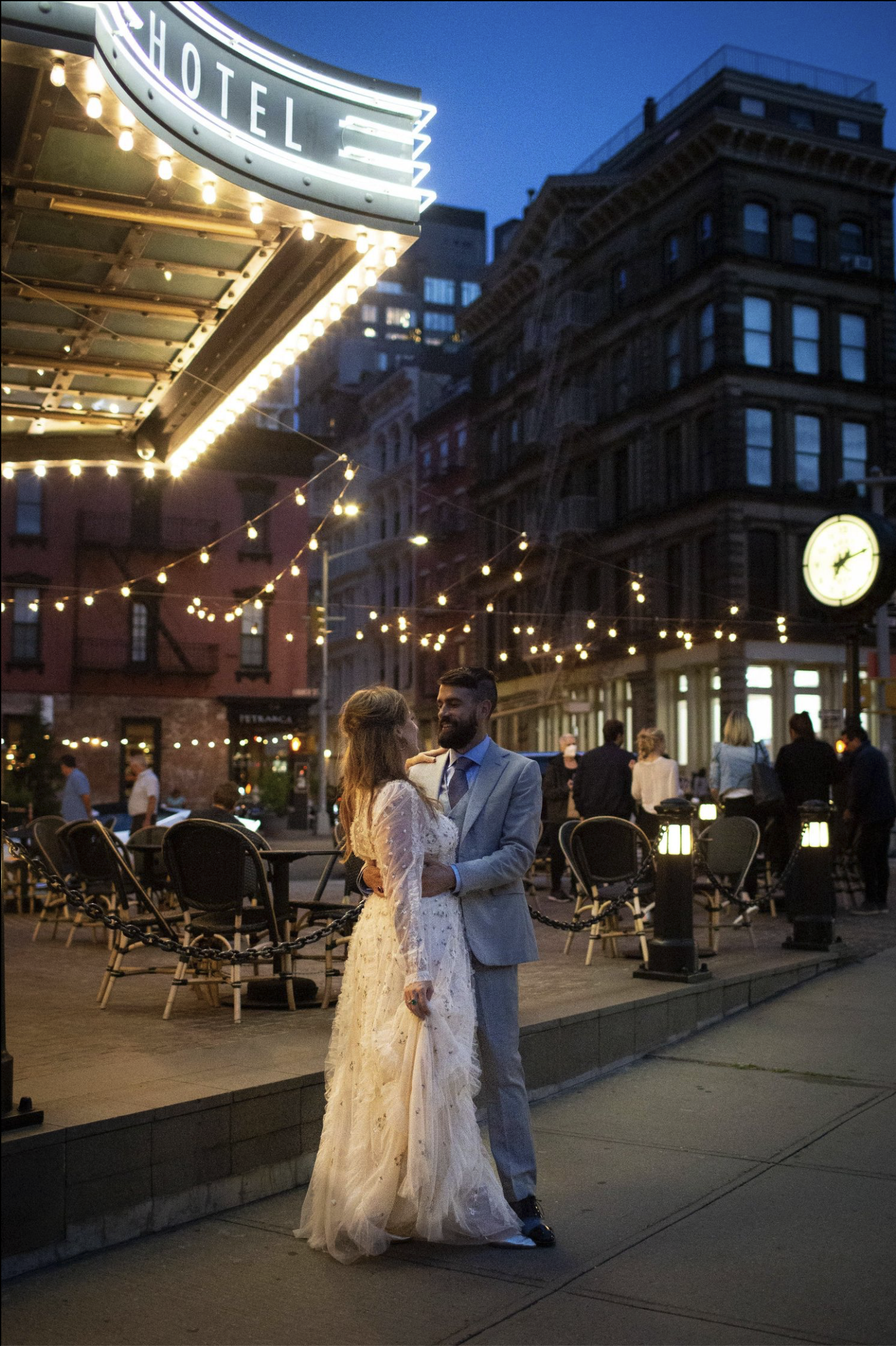 A newlywed couple dancing outside at night, with string lights overhead and a city street background, including a hotel marquee and a clock on a building.