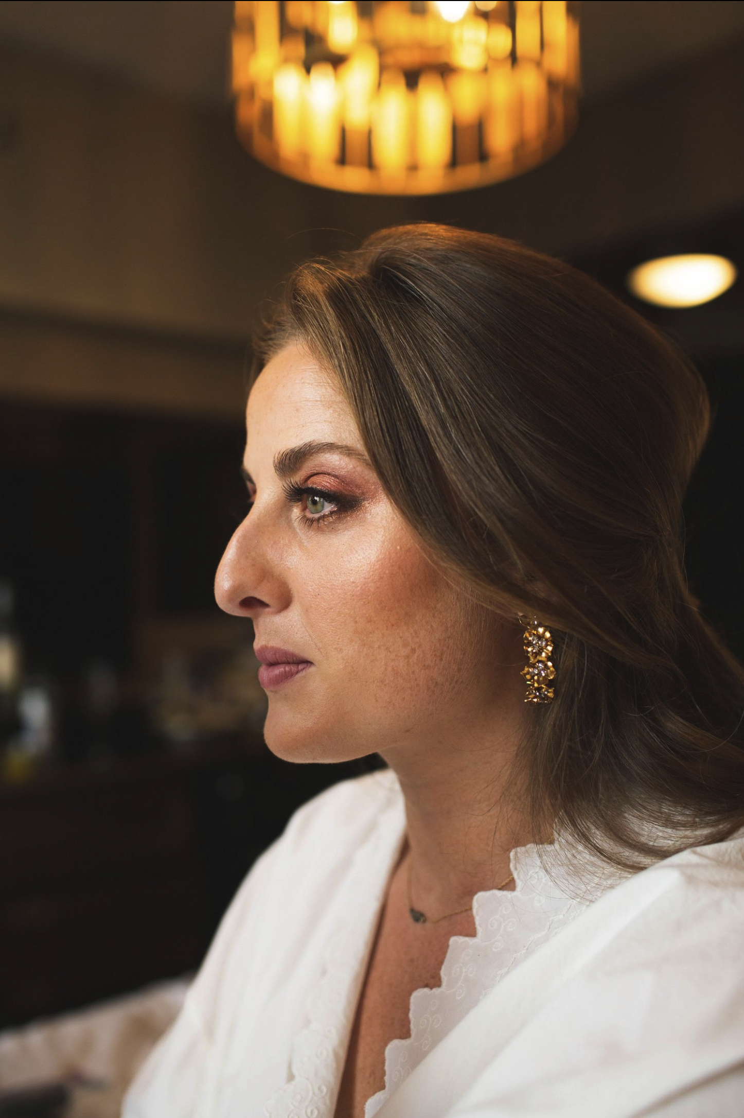 Close-up of a woman with brown hair styled in soft waves, wearing gold earrings and a white top with scalloped edges, looking to her left in a dimly lit room with warm lighting.