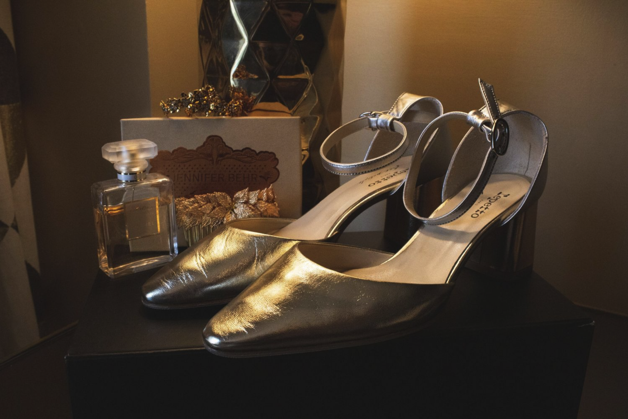 A pair of metallic gold high-heeled shoes on a black display surface, with a clear perfume bottle, a jewelry box, and a decorative box labeled 'Jennifer Behr' behind them.