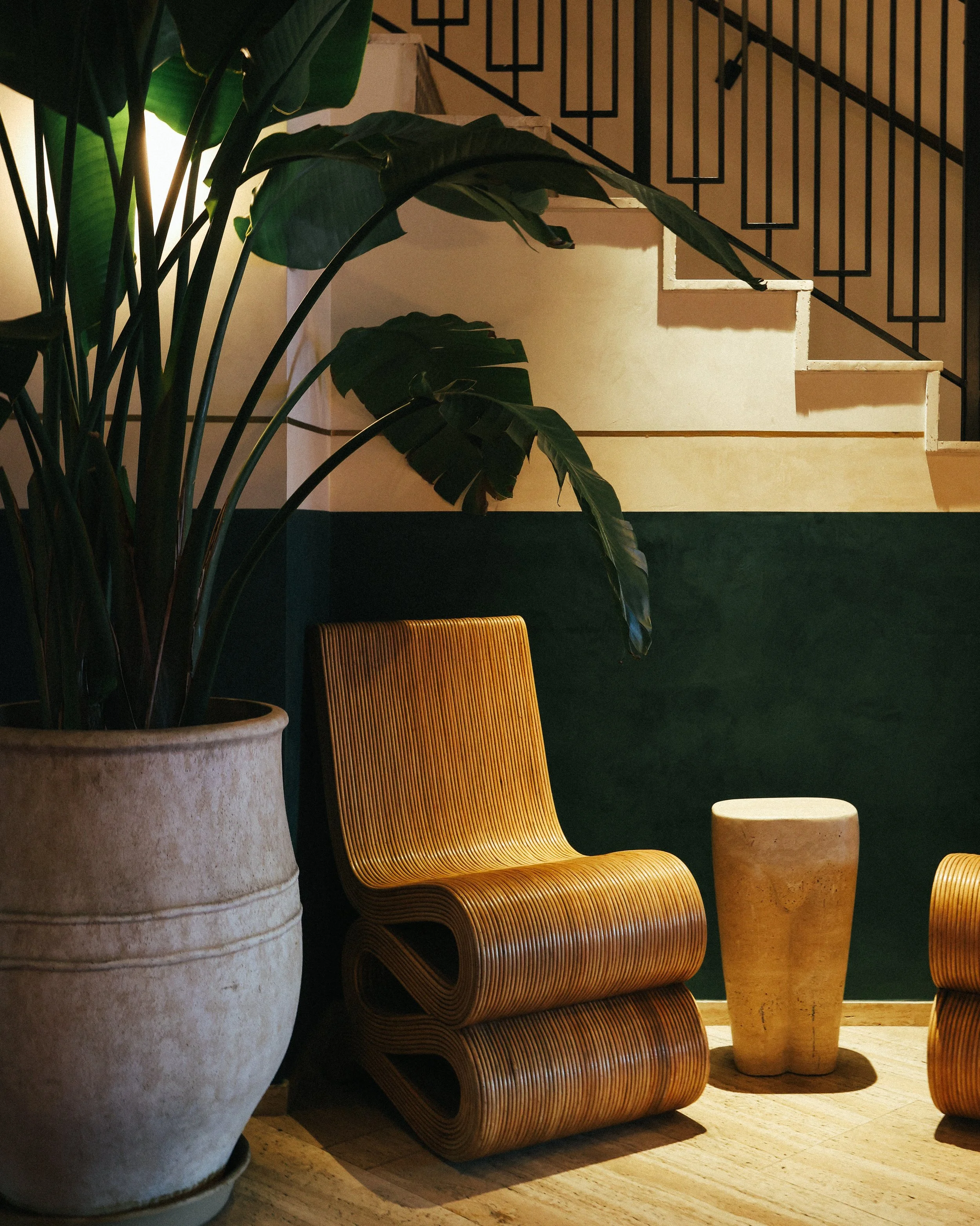 Interior scene featuring a large potted plant with broad green leaves, a modern wooden chair with a curved design, a round stone side table, and a staircase with black metal railing in the background, illuminated by warm lighting.