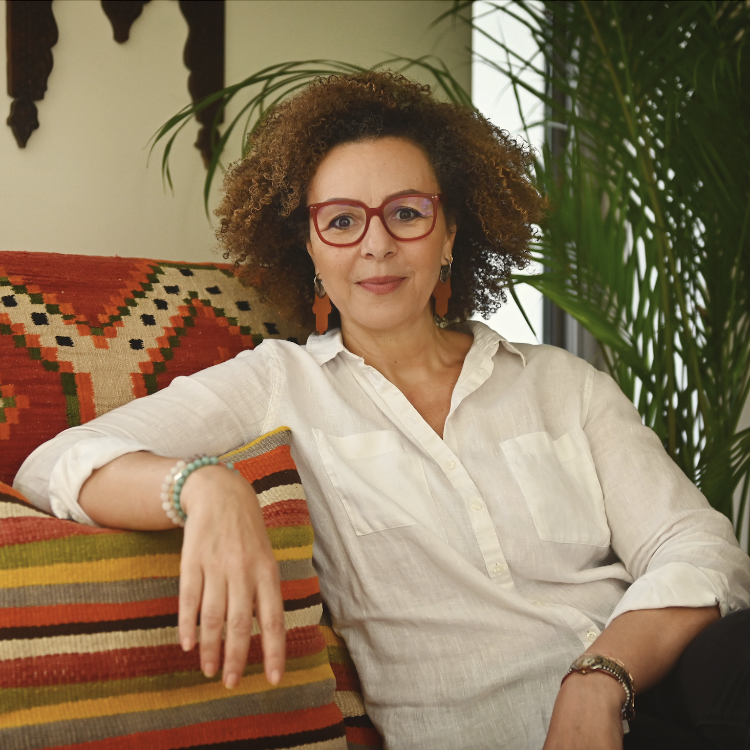 A woman with curly hair and red glasses sitting on a colorful striped couch with a green plant behind her, smiling at the camera.