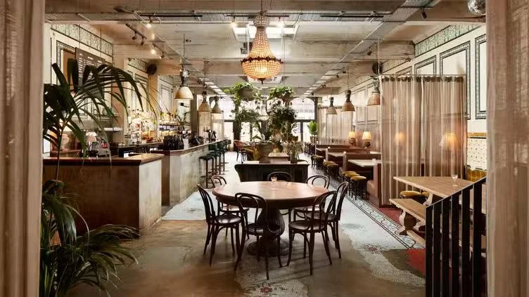 Interior of a cozy, stylish cafe with round table and chairs, plants, warm lighting, and decorative curtains.
