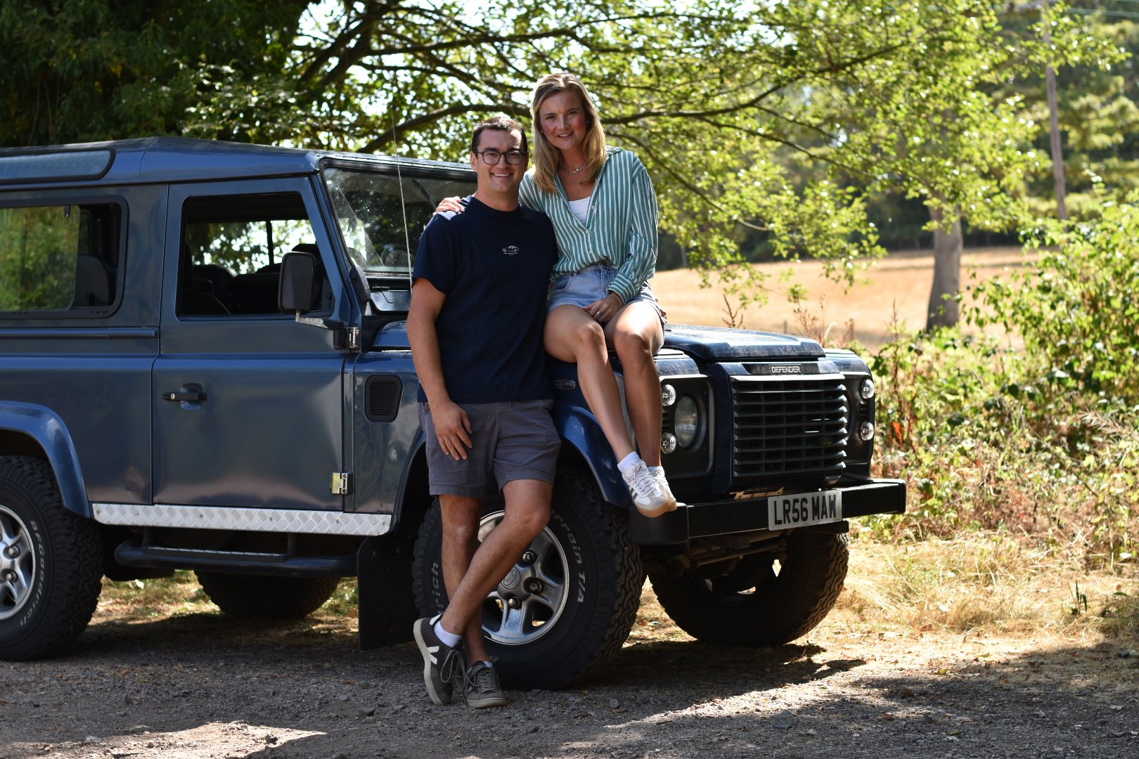 A smiling young man and a young woman sitting on the hood of a blue Land Rover Defender in a wooded area on a sunny day.
