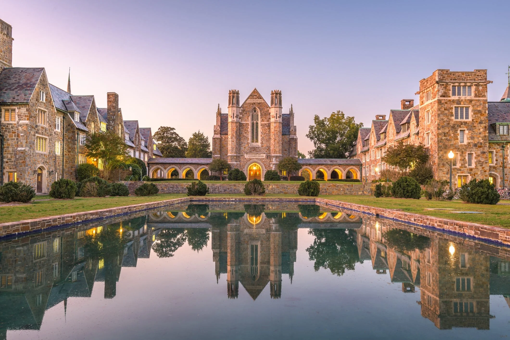 A college campus of stone buildings and a chapel, reflected in a long pool at dusk.