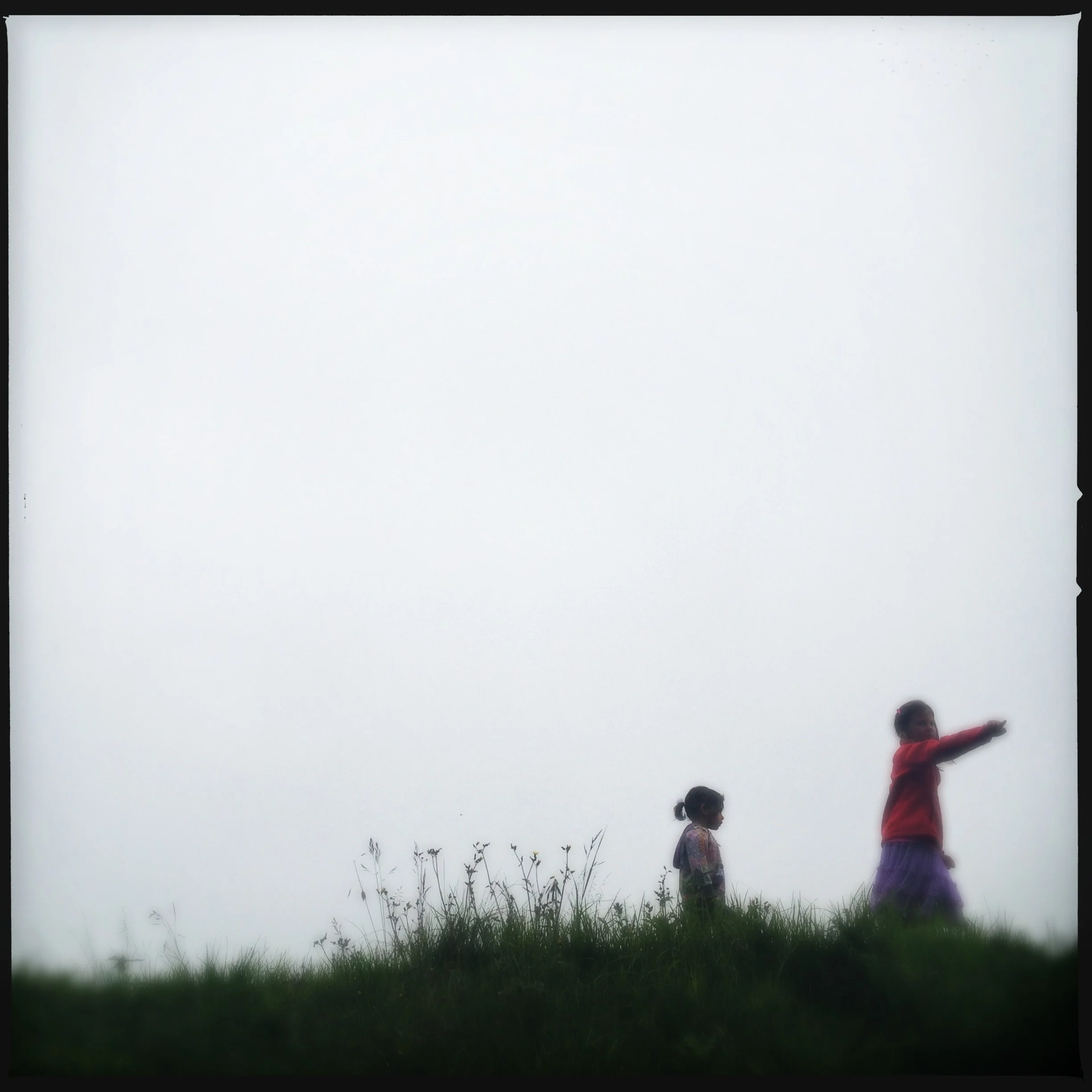 Two small figures, a child and an adult, on a grassy hilltop under a wide, foggy sky.