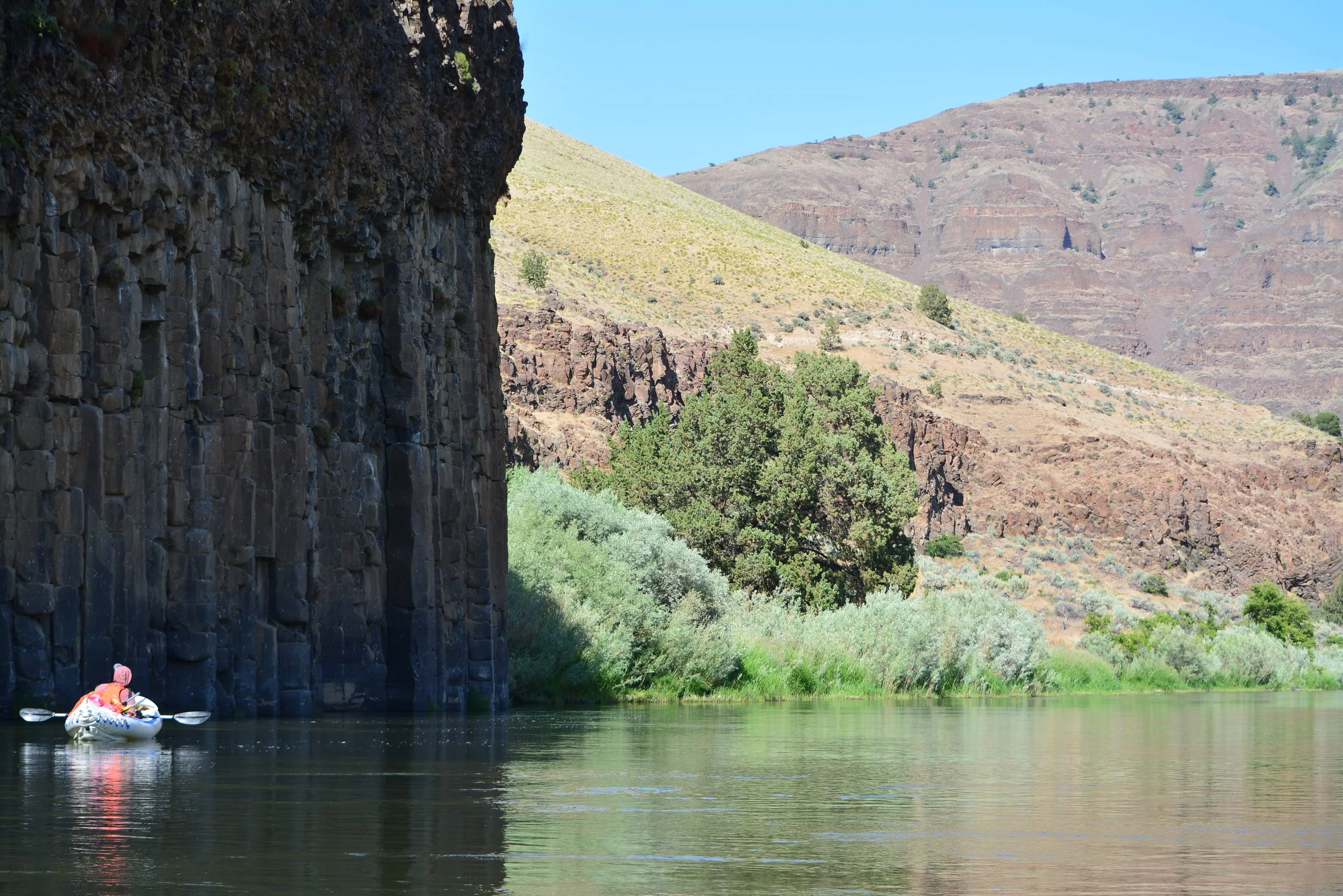 A person kayaking on a calm river near tall dark rock formations and arid hills under a clear blue sky.
