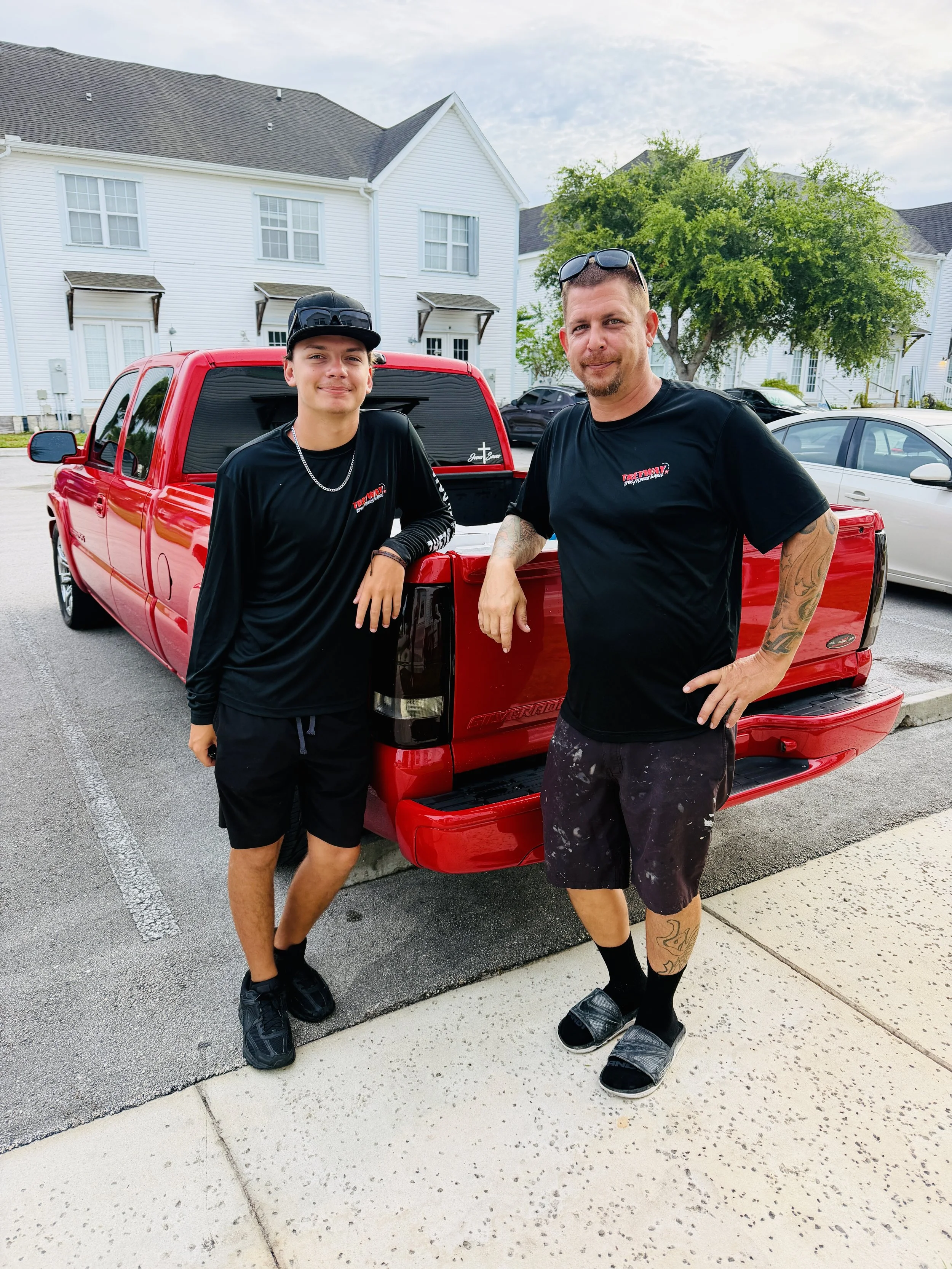 Two men standing next to a red pickup truck parked on a suburban street. One younger man with a black cap, chain necklace, and black shirt, and an older man with sunglasses on his head, tattoos, and black shirt with red and white lettering.
