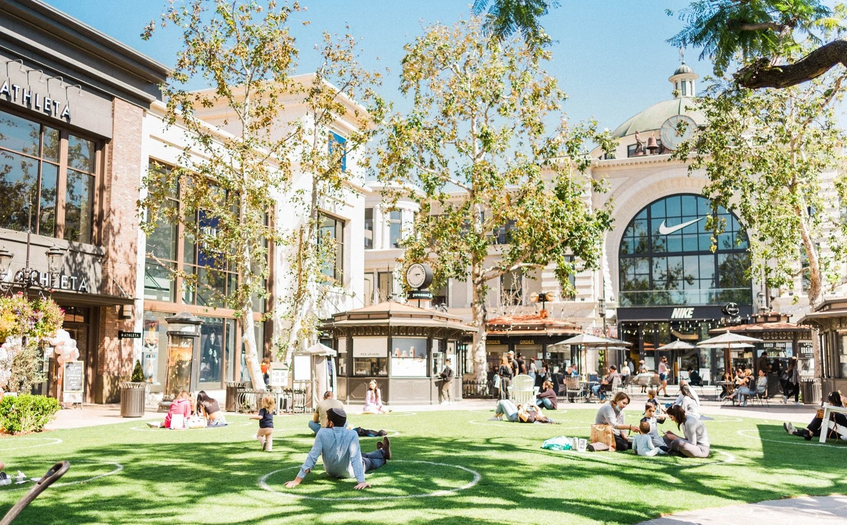 A bustling outdoor shopping plaza with people sitting on the grass and walking around, shops including Nike and Athleta, trees with green leaves, and a bright, sunny sky.