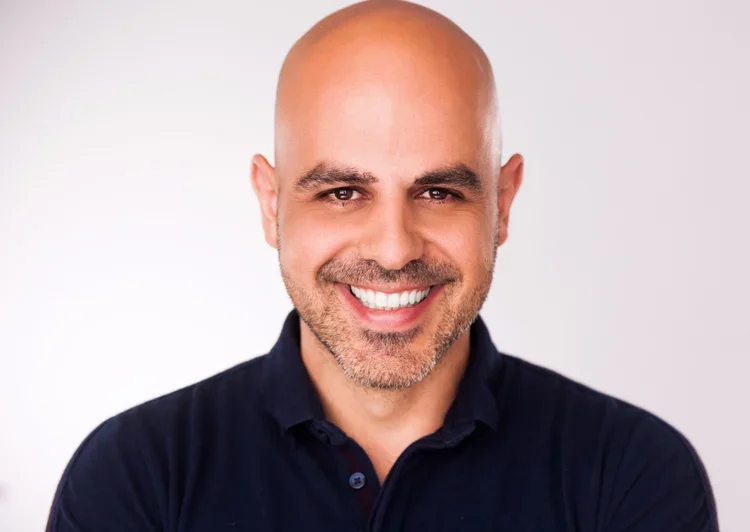 Headshot of smiling bald man with a beard, wearing a navy blue collared shirt against a light background.