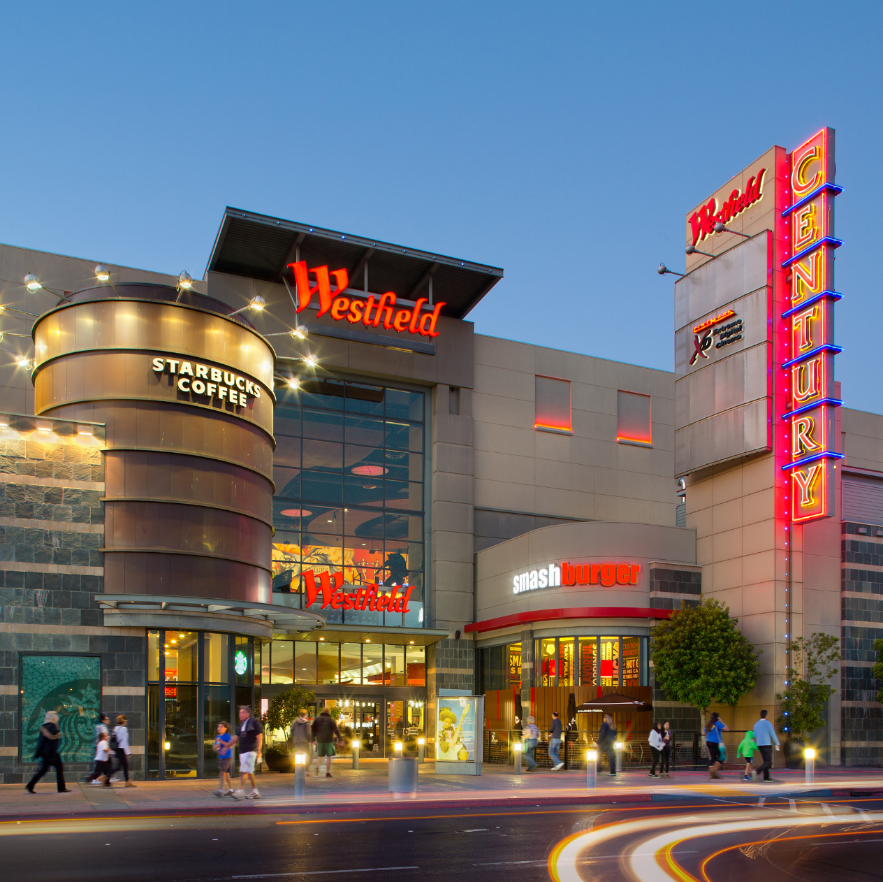 Exterior of Westfield shopping mall at dusk with neon signage, Starbucks Coffee, Smashburger, and multiple store signs, with people walking on the sidewalk.
