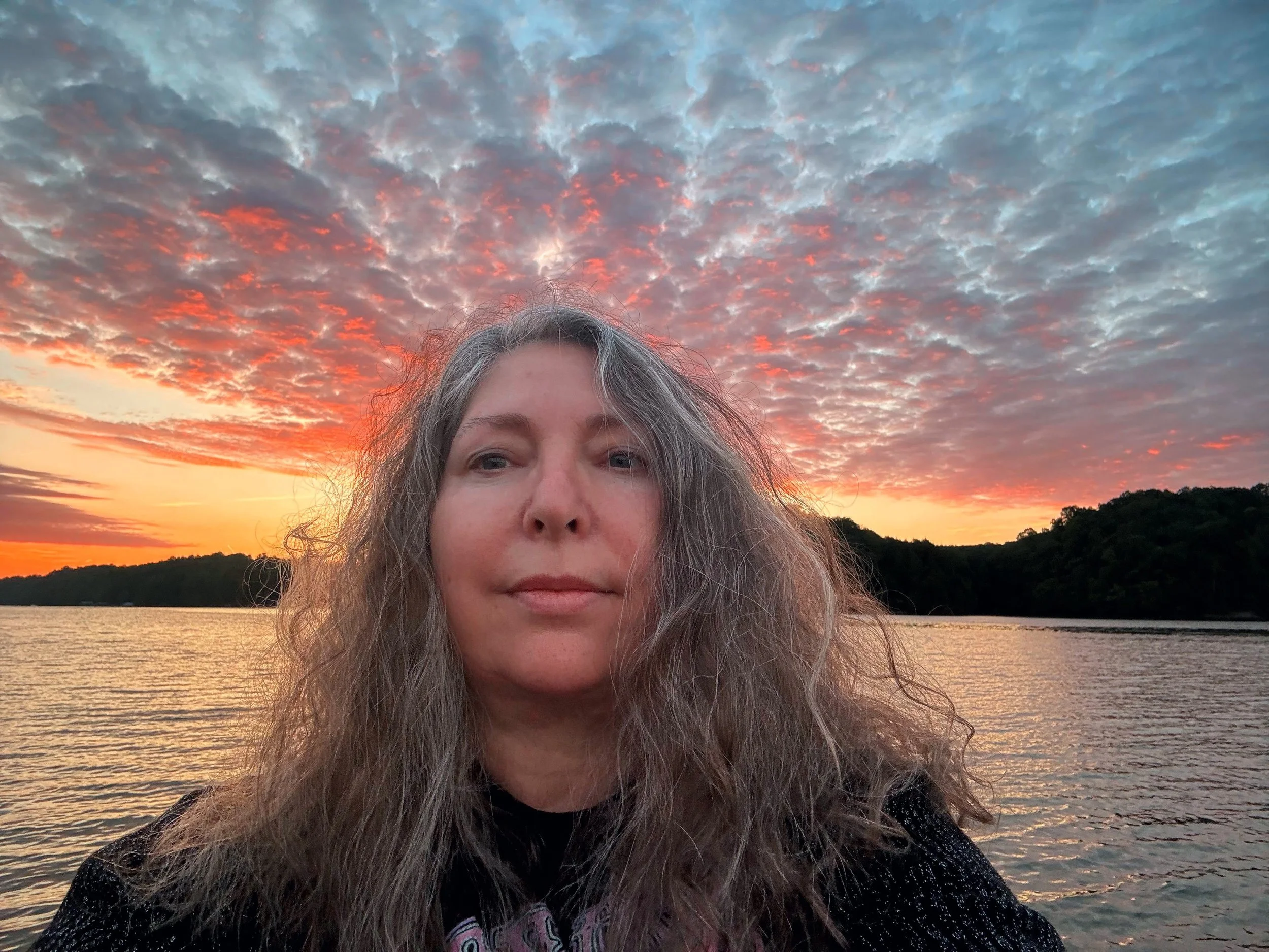 A woman with long gray hair taking a selfie by a body of water during a vibrant sunset with an orange and pink sky filled with clouds.