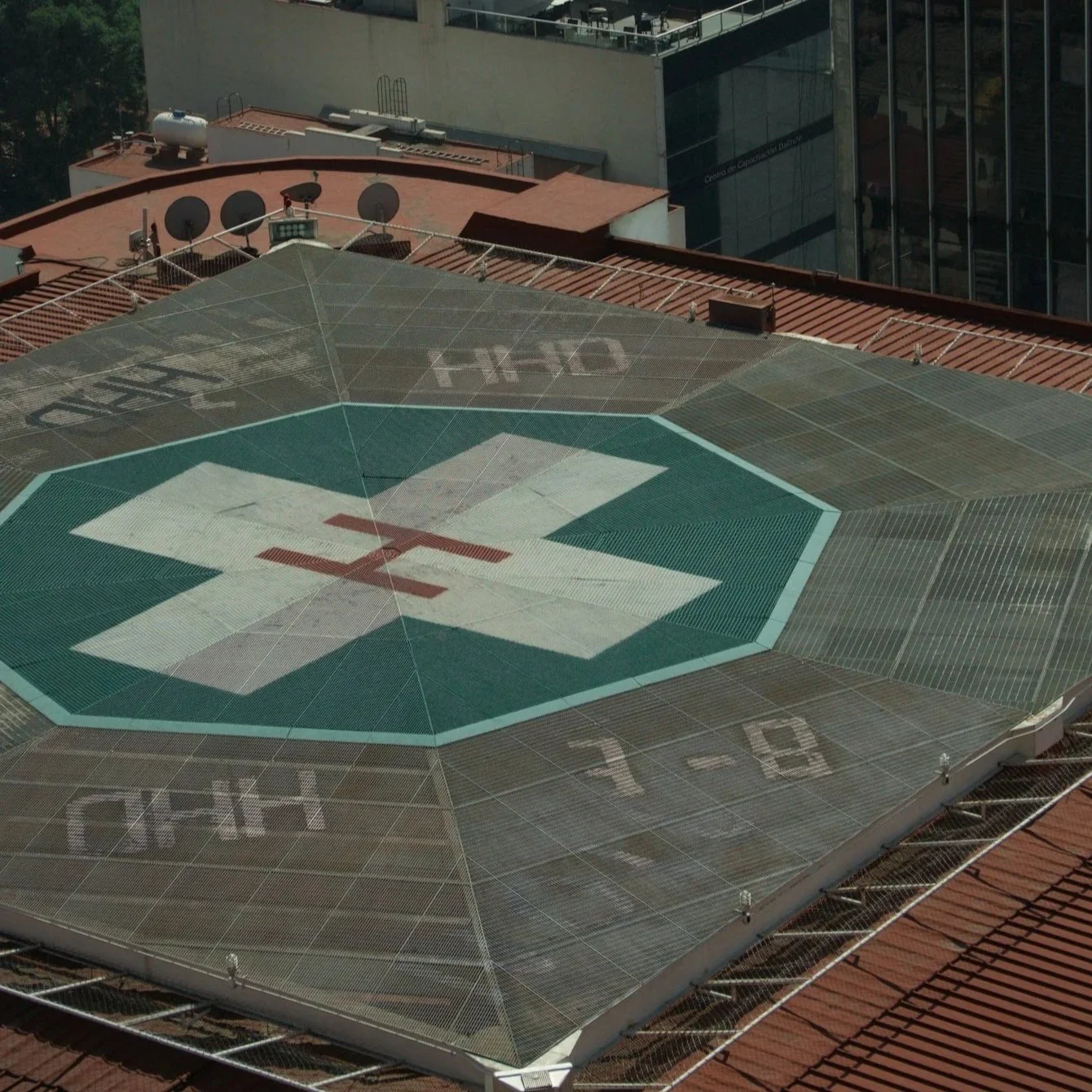 View of a rooftop featuring a helipad with a green and white cross symbol and a red 'H', and surrounding buildings with satellite dishes and an office building with glass windows.