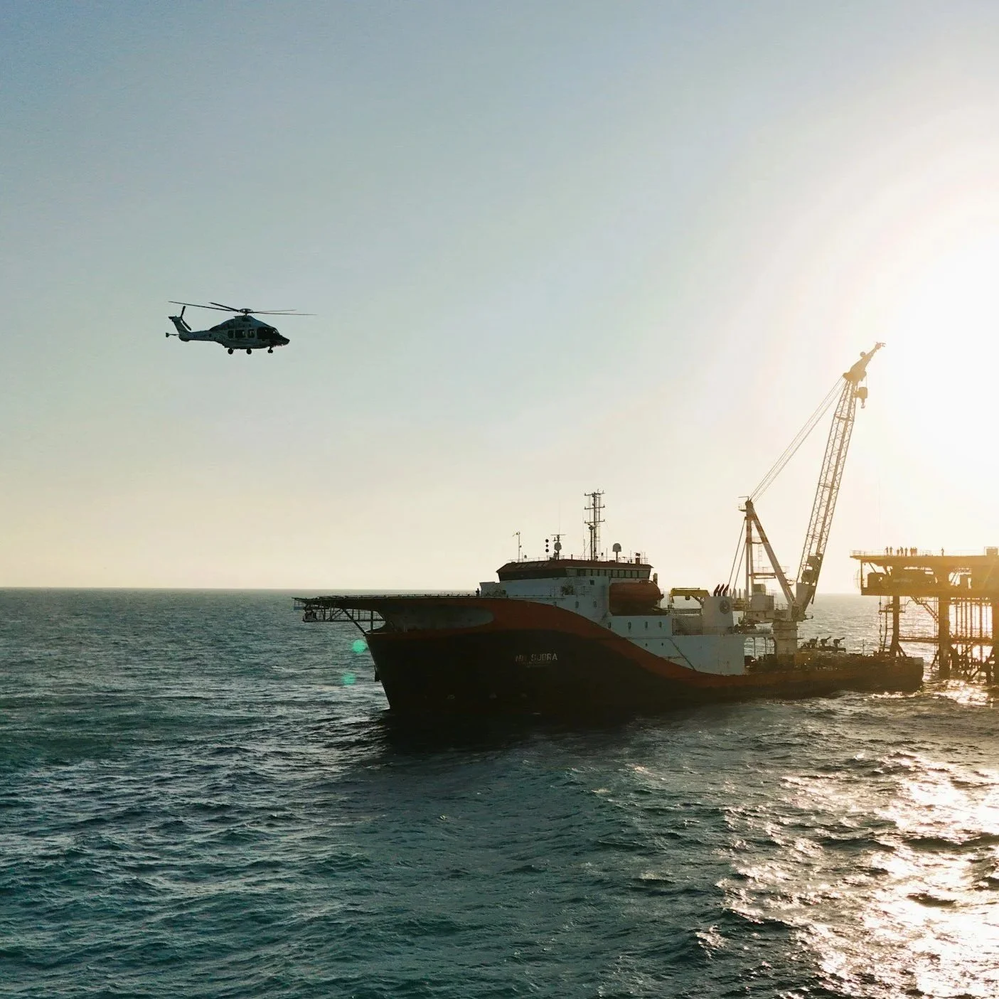 A ship at sea with a helicopter flying nearby against the backdrop of the sun setting or rising.