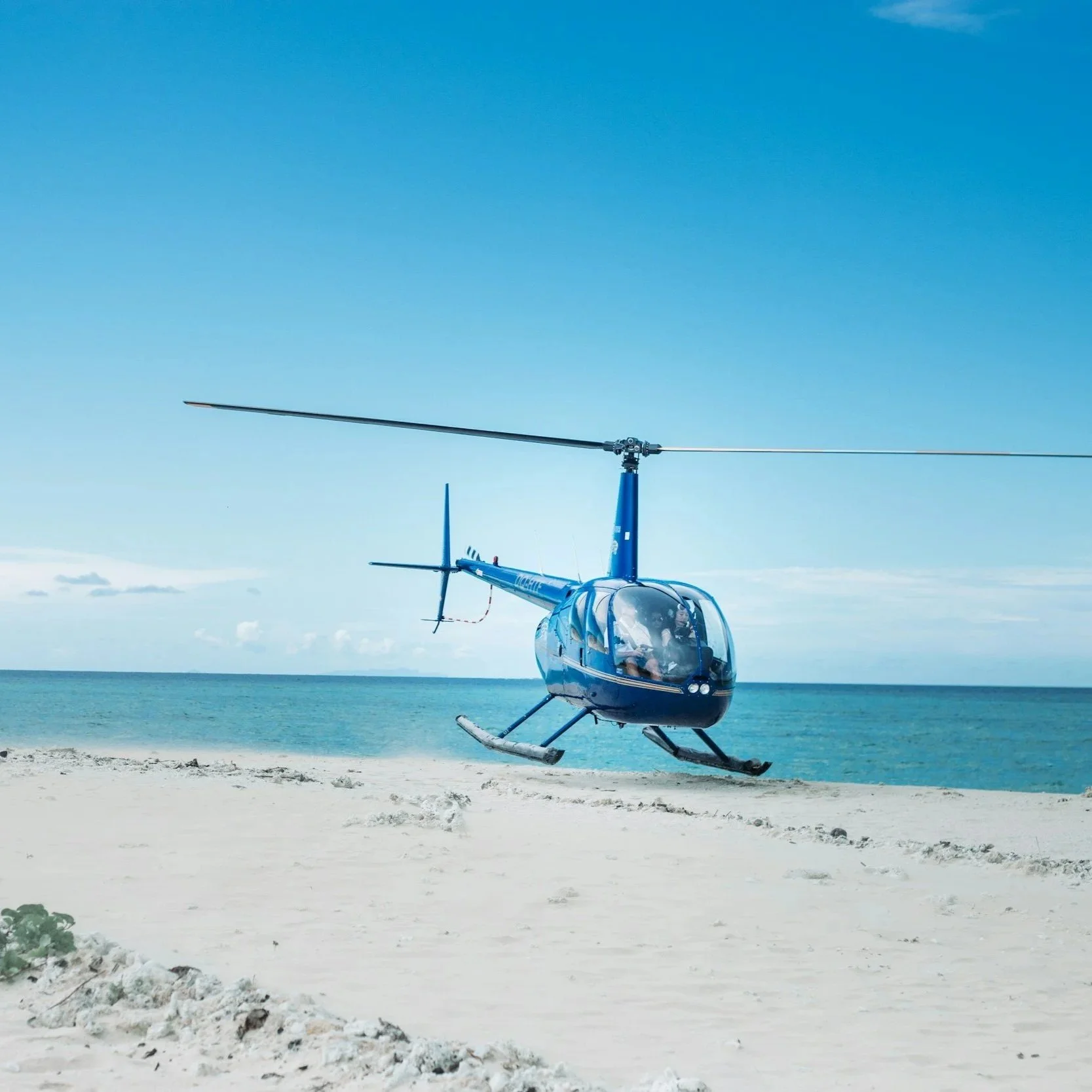 Blue helicopter hovering above a sandy beach with turquoise water and blue sky.