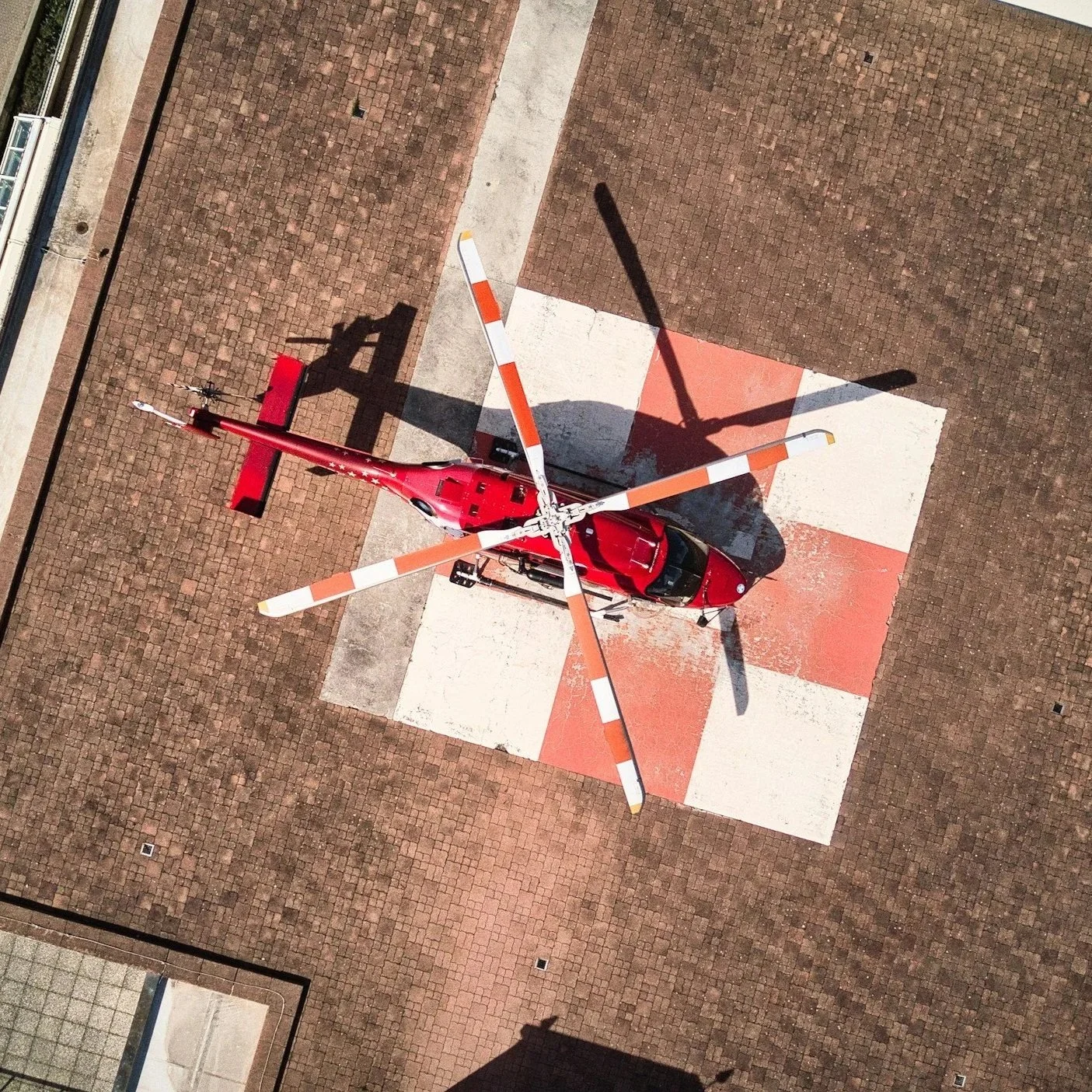 From above, a red helicopter is parked on a helipad with white and orange markings, surrounded by brick pavement.
