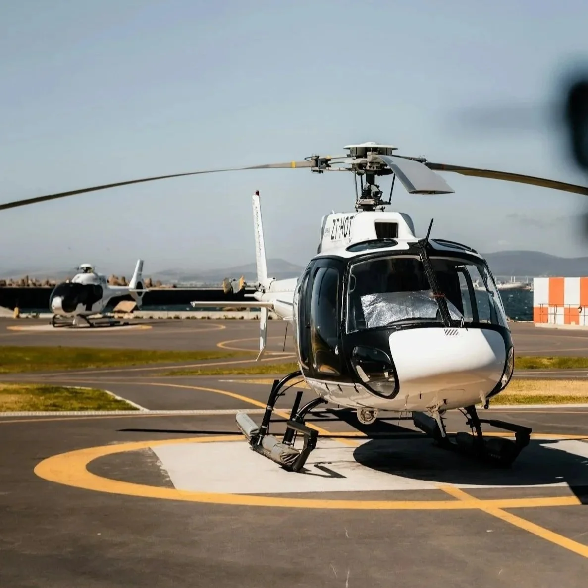 A white helicopter with black accents on a helipad, with another helicopter in the background and a landscape of mountains and sky.
